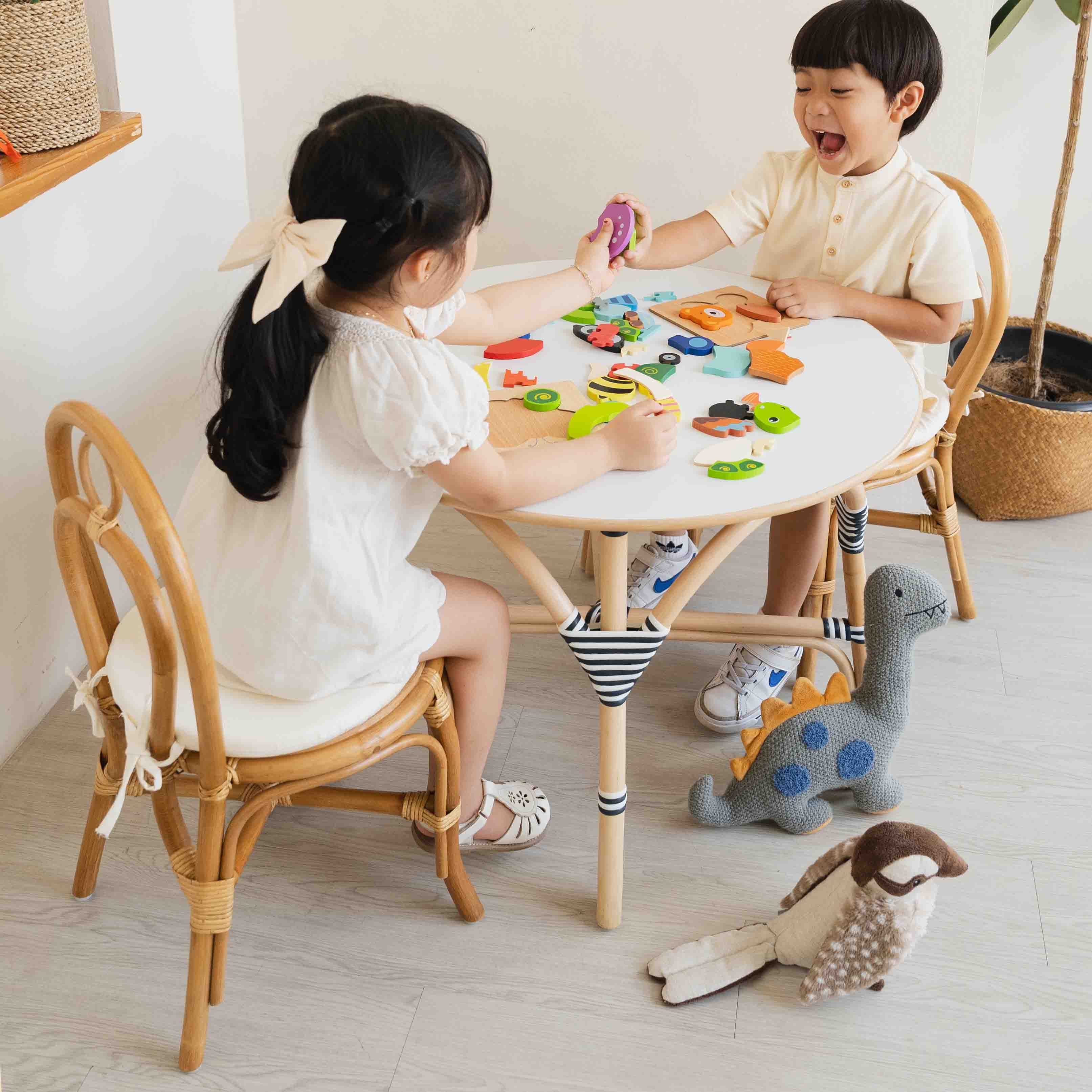 Two children sitting on handcrafted rattan wicker Amélie Kids Chairs by MOMIJI pair with the Aulia Kids Table, playing with colorful puzzle toys in a joyful indoor playtime.