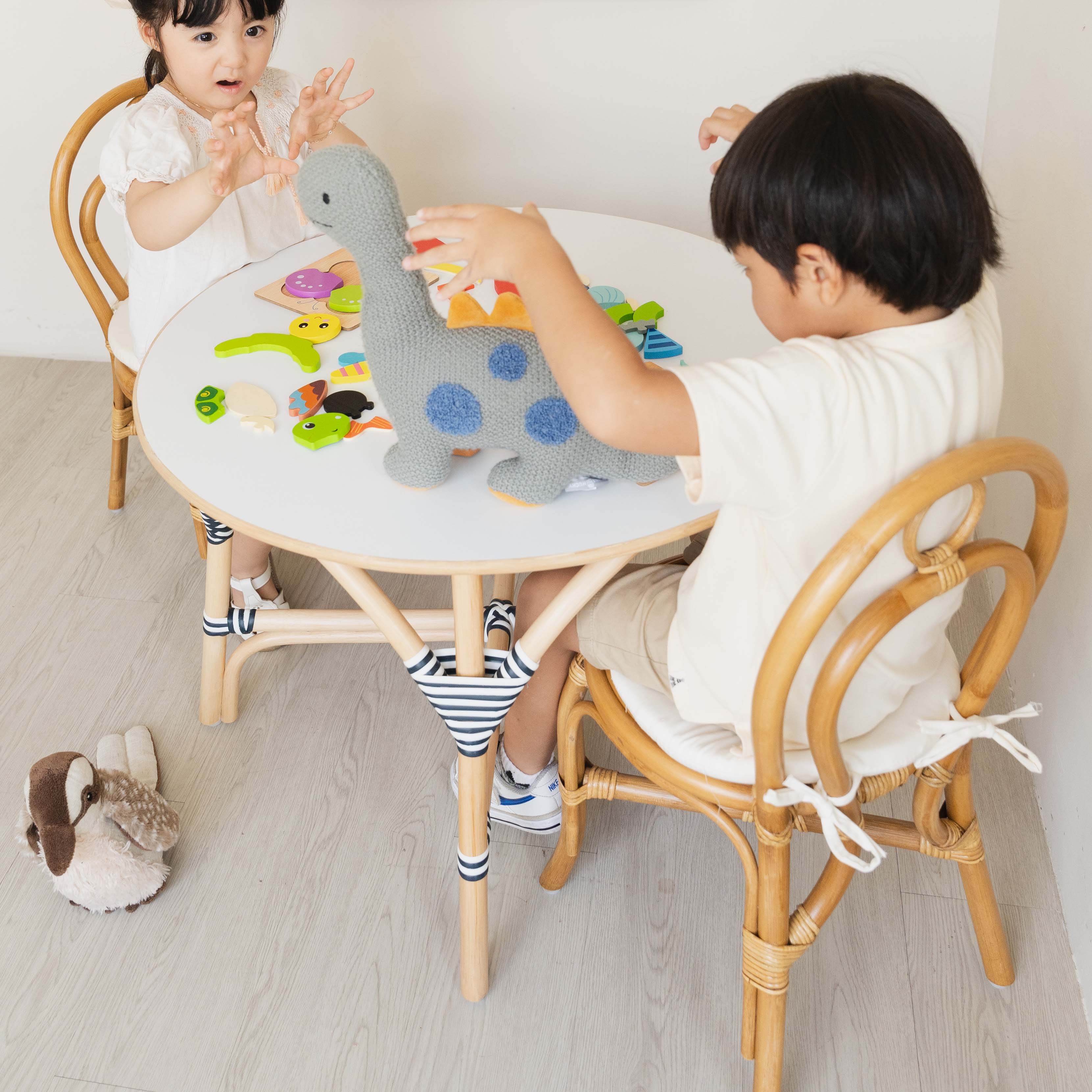 Two children sitting on the handcrafted rattan wicker Amélie Kids Chairs by MOMIJI pair  setwith the Aulia Kids Table, playing with a dinosaur toy on a round table in a bright indoor setting.