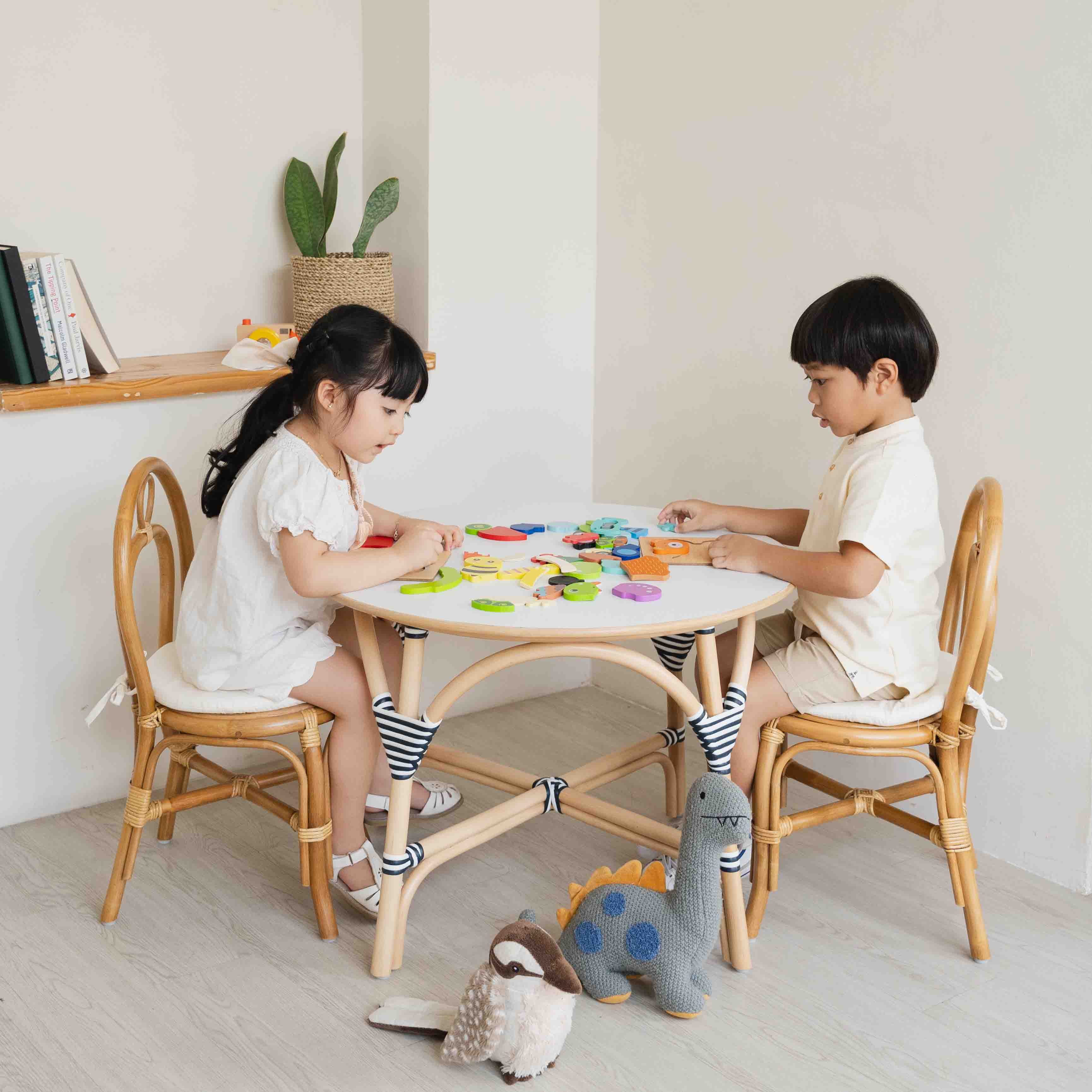 Two children sitting on the handcrafted rattan wicker Amélie Kids Chairs by MOMIJI at the Aulia Kids Table, playing with toys together at a small round kids table in a bright indoor setting