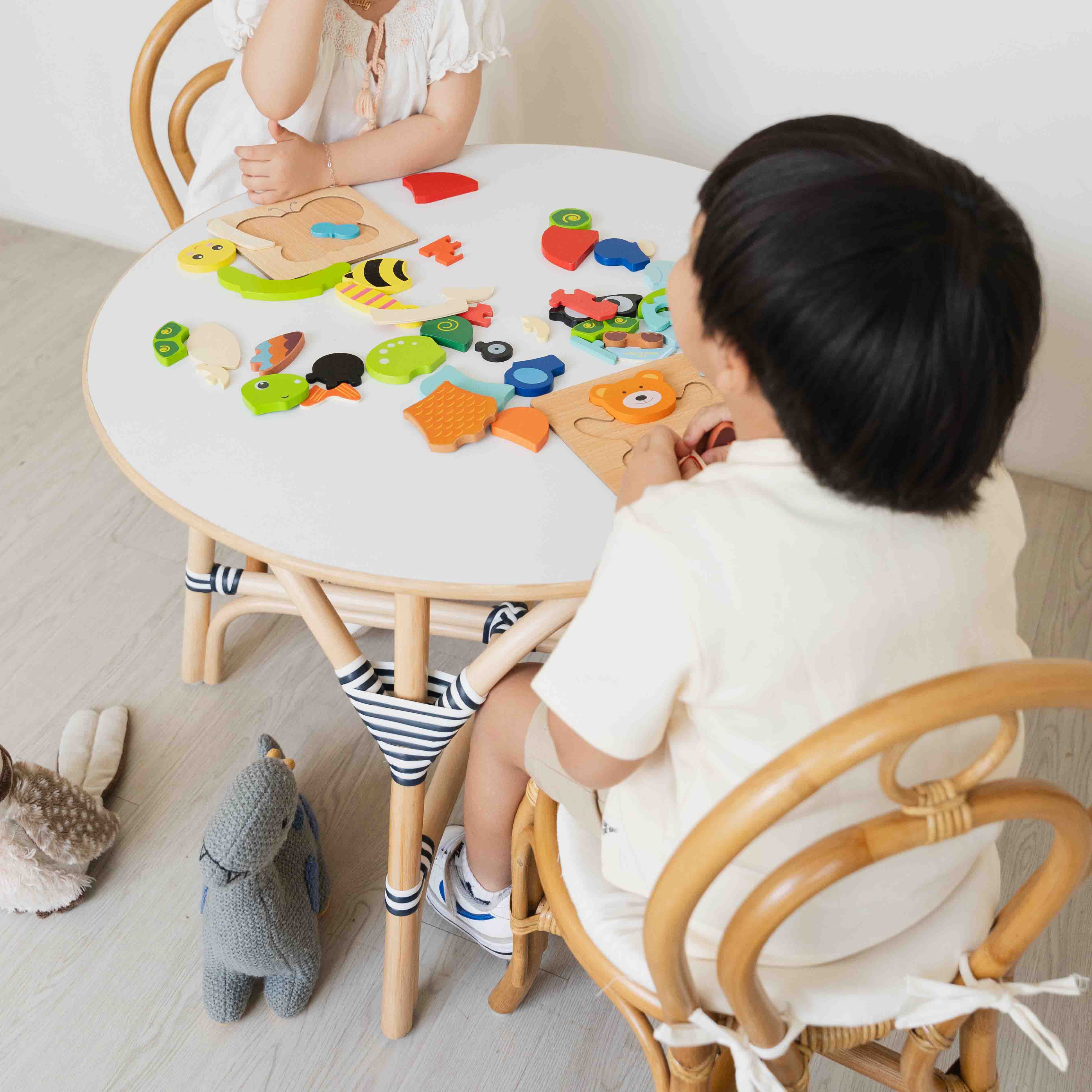 Top view of two children sitting on the handcrafted rattan wicker Amélie Kids Chairs by MOMIJI pair set with the Aulia Kids Table, playing with colorful puzzle toys, highlighting the sturdy navy-white binding details on the rattan structure and the smooth HPL-laminate tabletop surface.