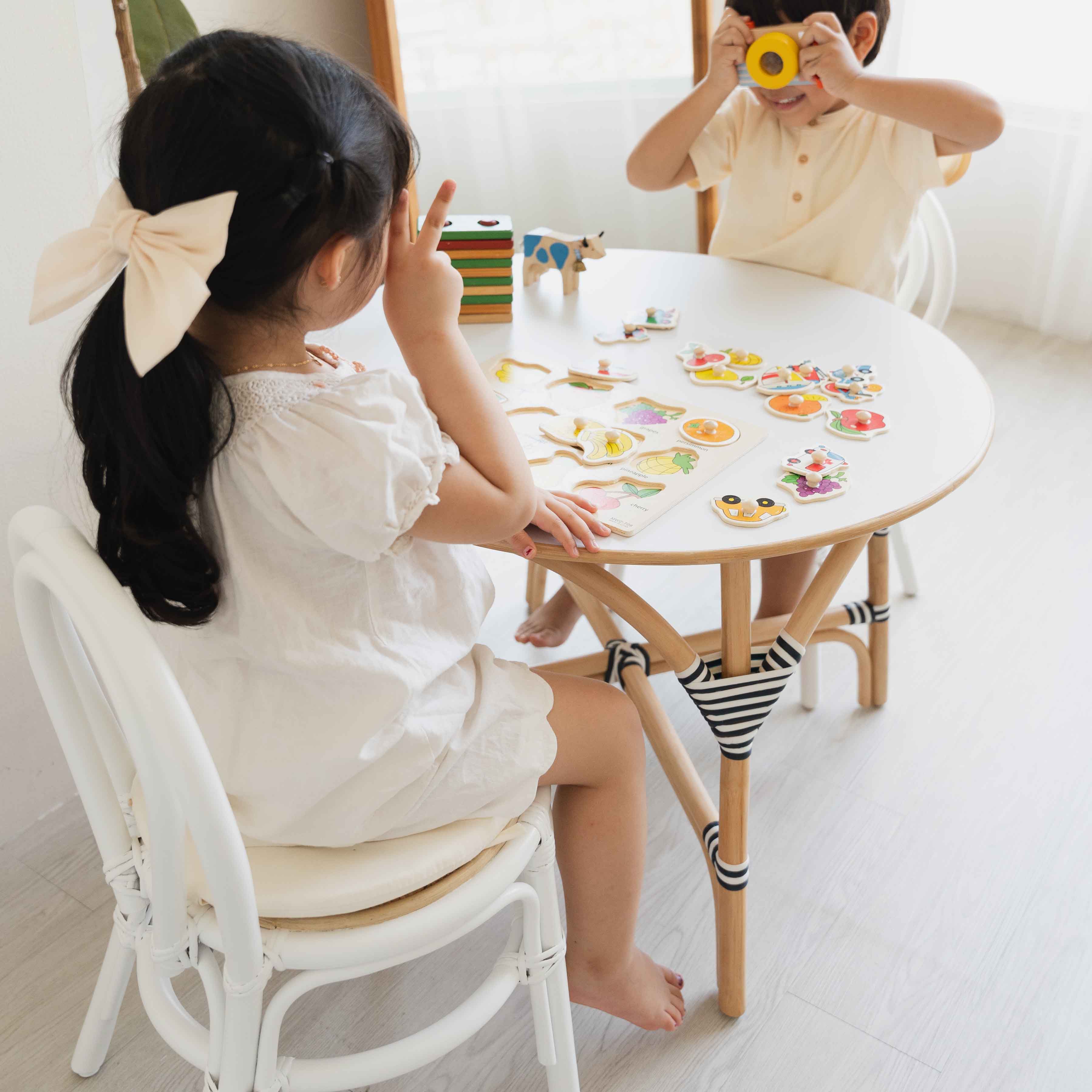 Two children sitting on Aulia Kids Chairs by MOMIJI while playing with a toy camera and colourful toys on an Aulia Kids Table in a bright, playful room.