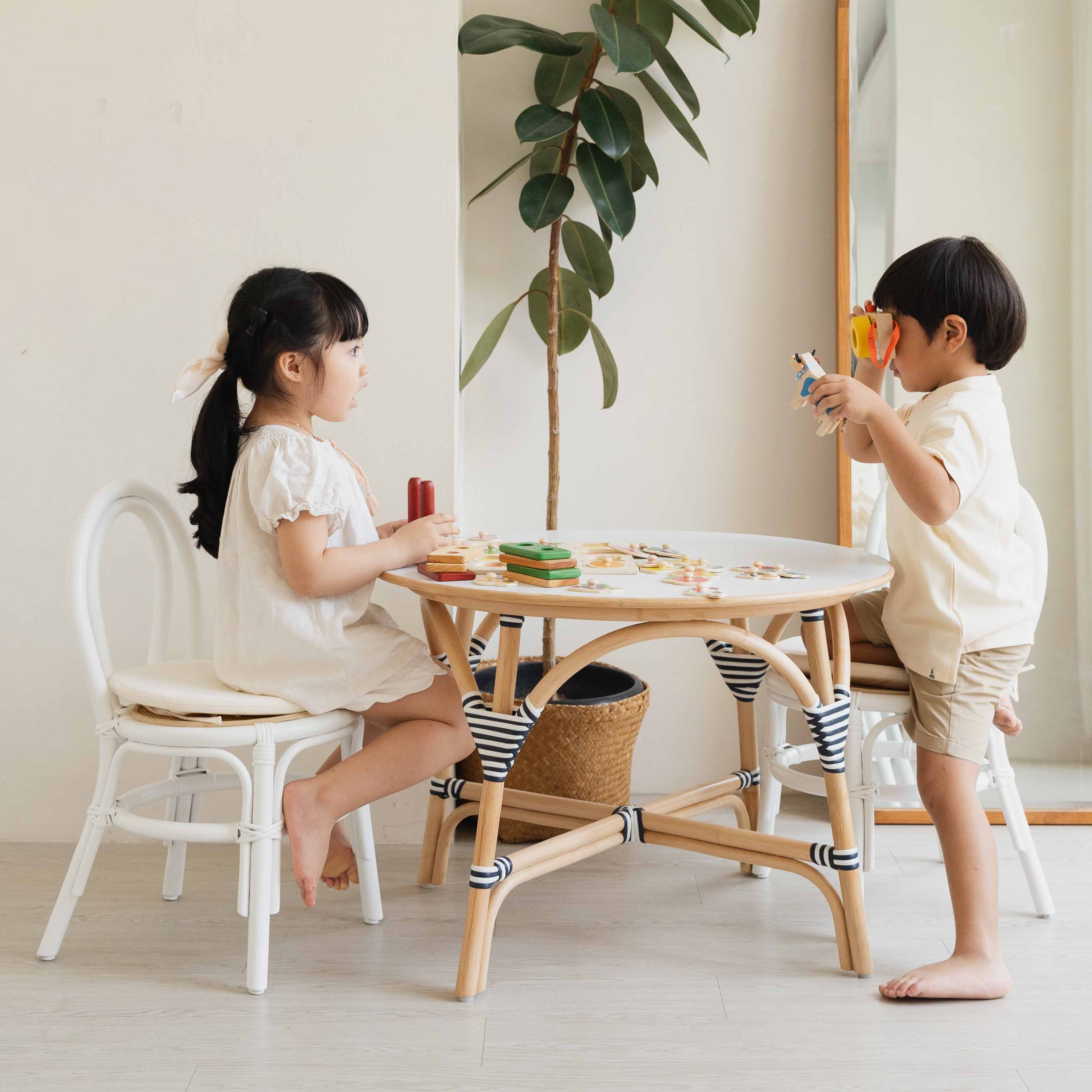 Two children sitting and playing together on Aulia Kids Chairs by MOMIJI, styled with toys on a handcrafted rattan Aulia Kids Table in a bright, playful room.
