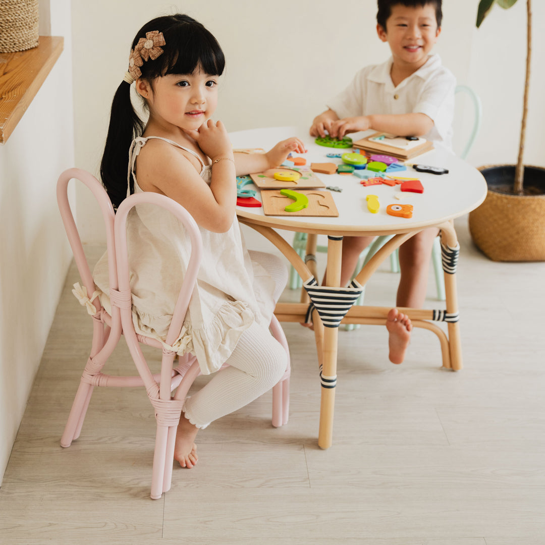 Two smiling children enjoying playtime on Pink and Mint Green handcrafted rattan wicker Bunny Chairs by MOMIJI at an Aulia Kids Table in a bright and playful children's room.