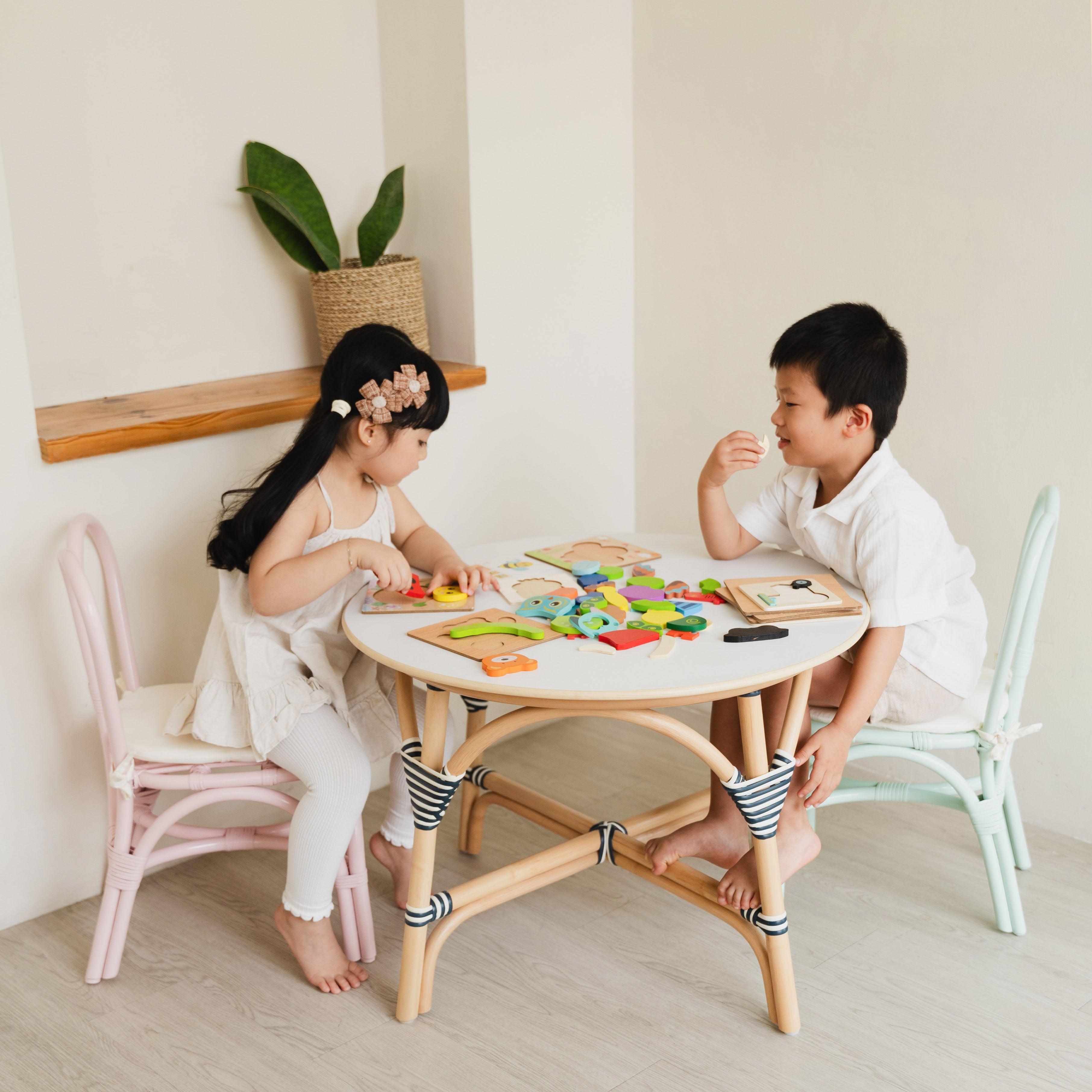 Two children sit on Pink and Mint Green Bunny Rattan Chairs by MOMIJI, playing with toys at an Aulia Kids Table in a bright and playful children's room.