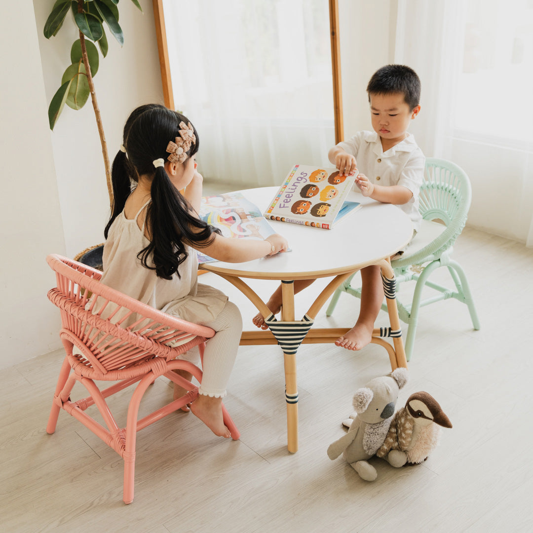 Two children sitting on Peach Pink and Mint Green Maya Kids Chairs by MOMIJI, reading books at an Aulia Kids Table in a room with a mirror and plants, with toys scattered on the floor.