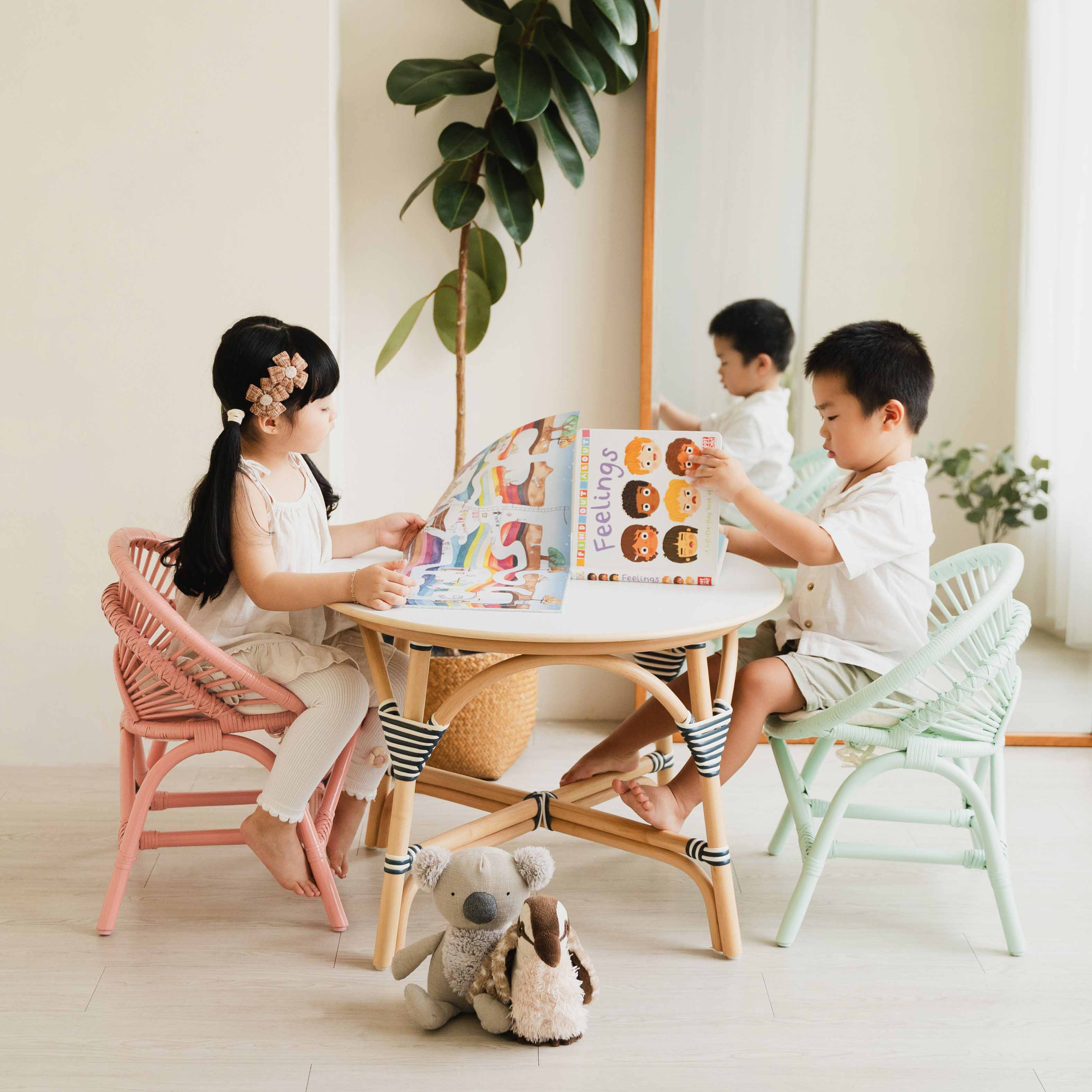 Two children sitting on on Peach Pink and Mint Green Maya Kids Chairs by MOMIJI, reading books at an Aulia Kids Table in a room with a mirror and plants.