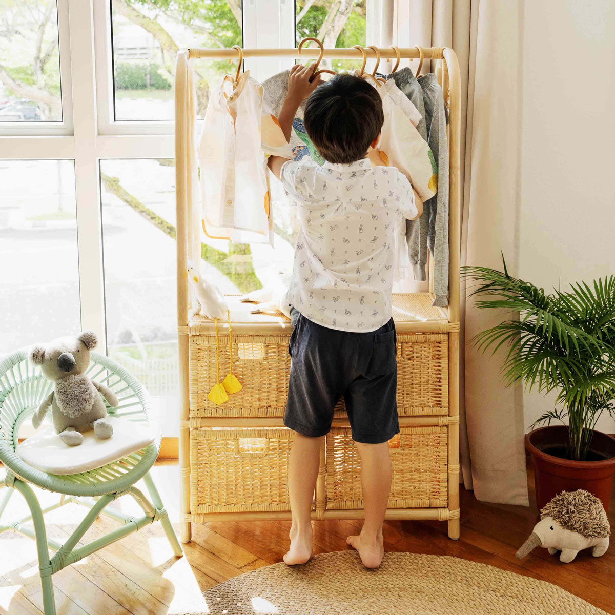 Child putting clothes in Belle Kids Clothing Rack with 4 Drawers by MOMIJI in a room with a window and plants.