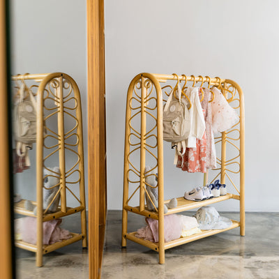 Natural handcrafted rattan Belle Kids Montessori Clothing Rack by MOMIJI with two built-in shelves, styled with baby clothes, backpacks, and shoes in a bright room, creating an organised and accessible storage space for children.