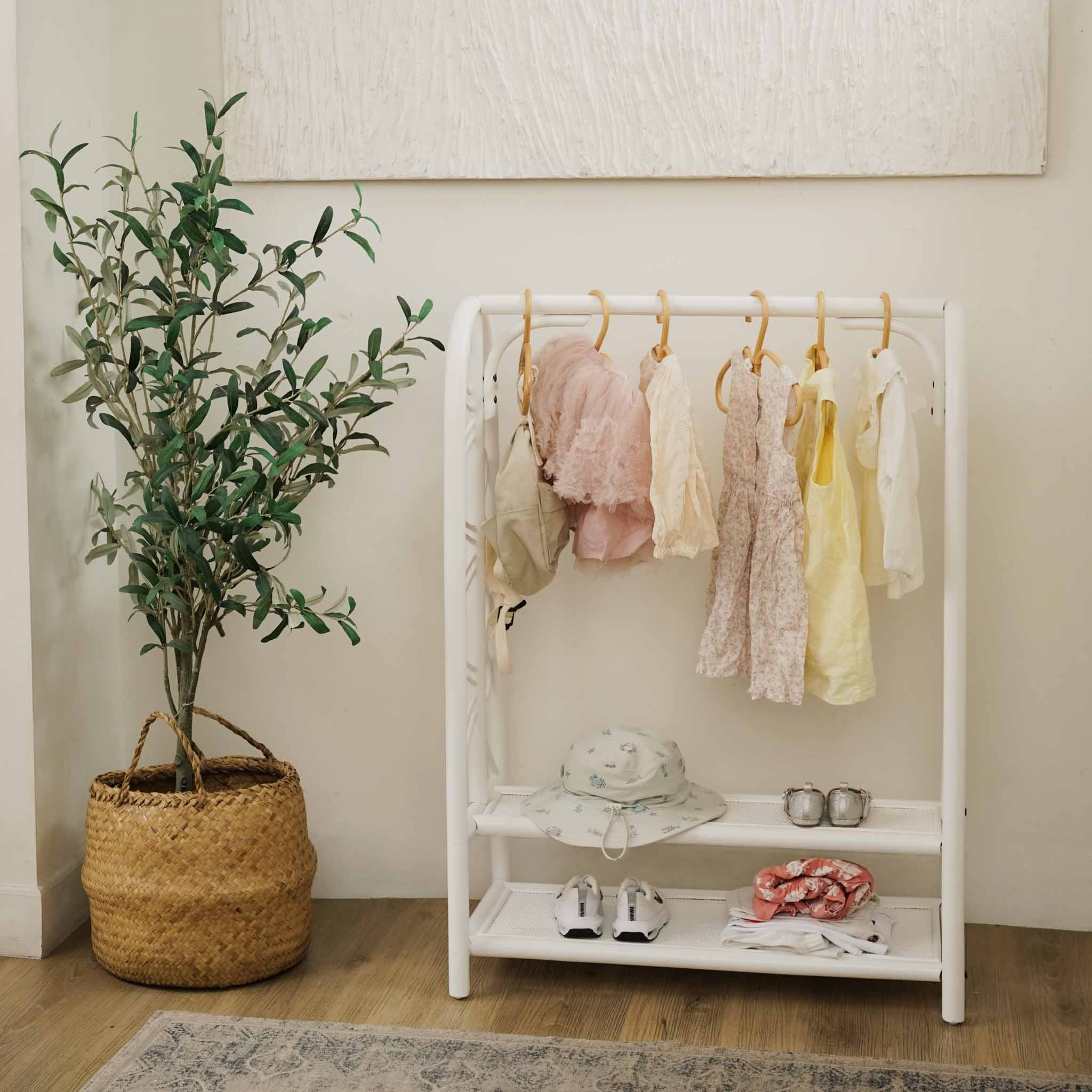 Handcrafted white rattan Belle Kids Montessori Clothing Rack by MOMIJI styled with baby clothes, a hat, shoes, and a small bag in a nursery-inspired setting beside a potted tree.