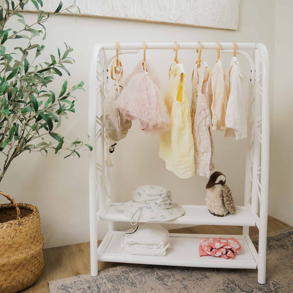 Handcrafted white rattan Belle Kids Montessori Clothing Rack by MOMIJI featuring a hanging rail and two storage shelves, styled with baby garments, a sun hat, a plush toy, and folded textiles in a nursery-inspired room.