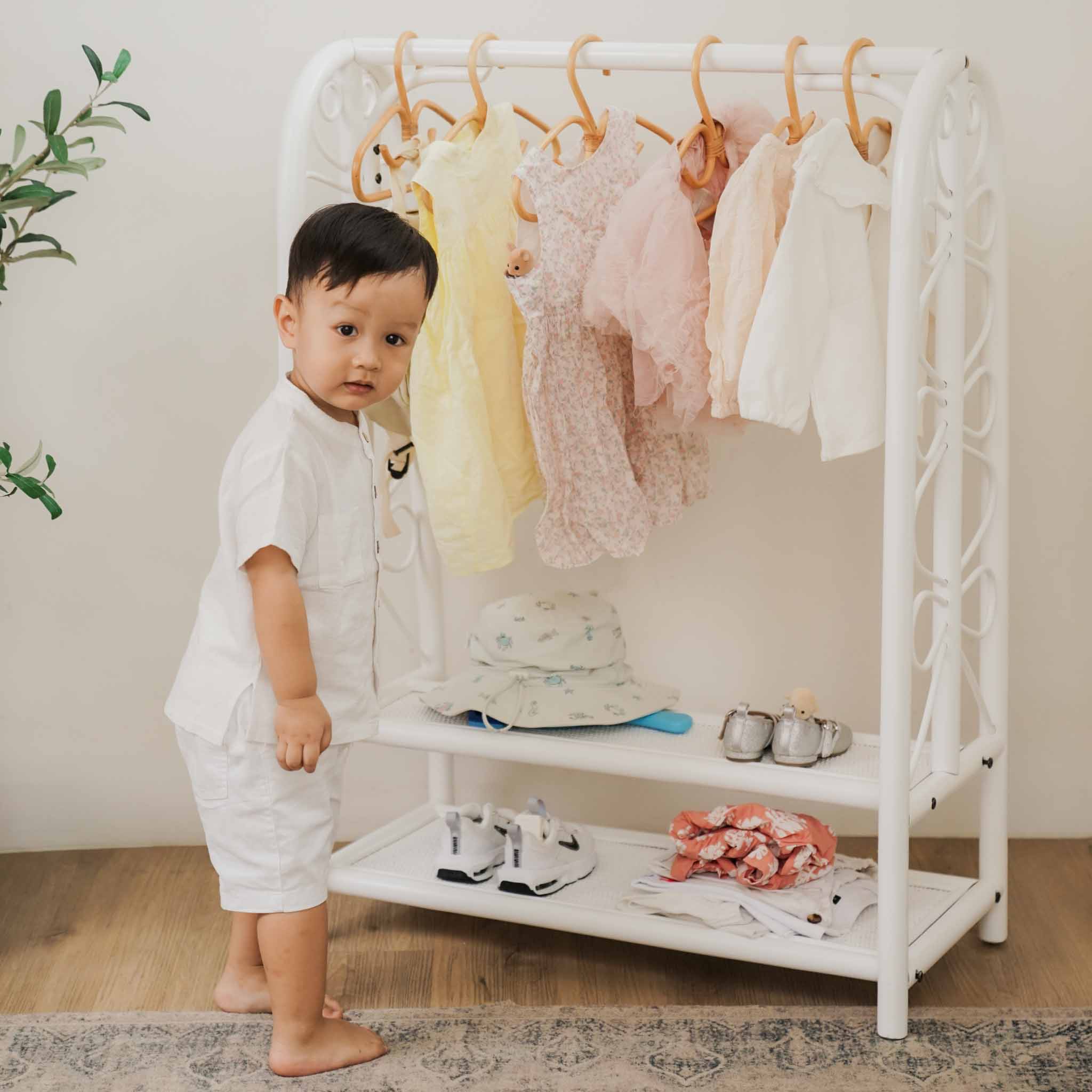 A toddler standing beside the handcrafted white rattan Belle Kids Montessori Clothing Rack by MOMIJI, featuring a child-friendly hanging rail and two lower shelves for storing shoes and accessories.