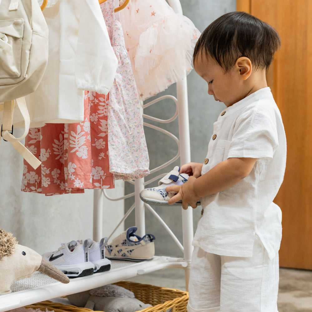 A toddler standing beside the handcrafted white rattan Belle Kids Montessori Clothing Rack by MOMIJI, holding a shoe while additional shoes are neatly stored on the lower shelves, showcasing multifunctional storage for children's essentials.