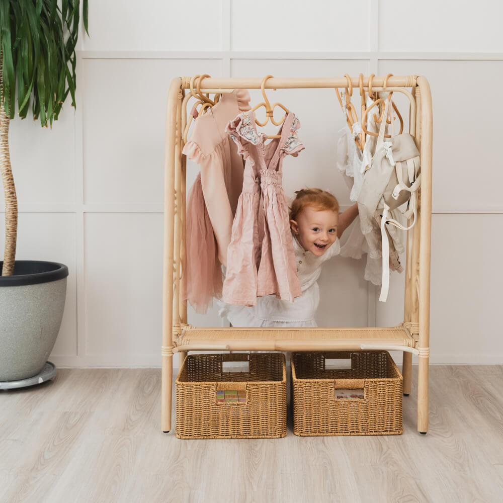 Child peeking from behind the Belle Kids Montessori Clothing Rack by MOMIJI with dresses hanging and extra rattan baskets on the floor.