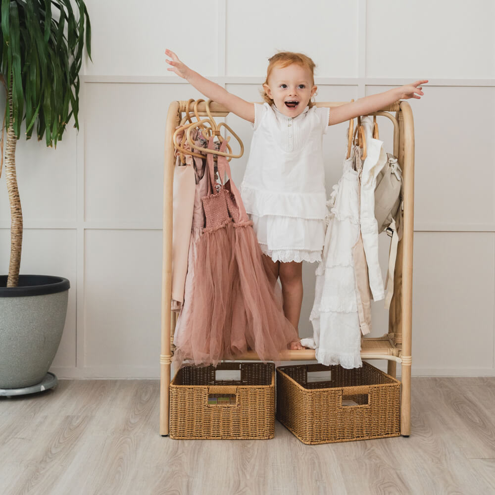 Child standing on the Belle Kids Montessori Clothing Rack by MOMIJI with dresses hanging and extra rattan baskets nearby.