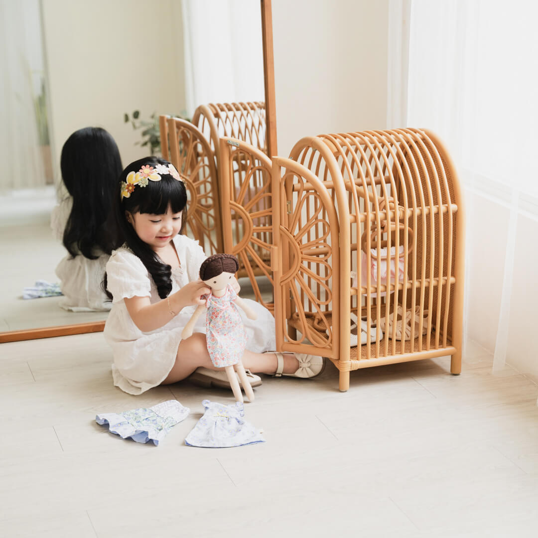 Child sitting on a wooden floor playing dress-up with dolls beside the Blossom Doll Closet by MOMIJI.