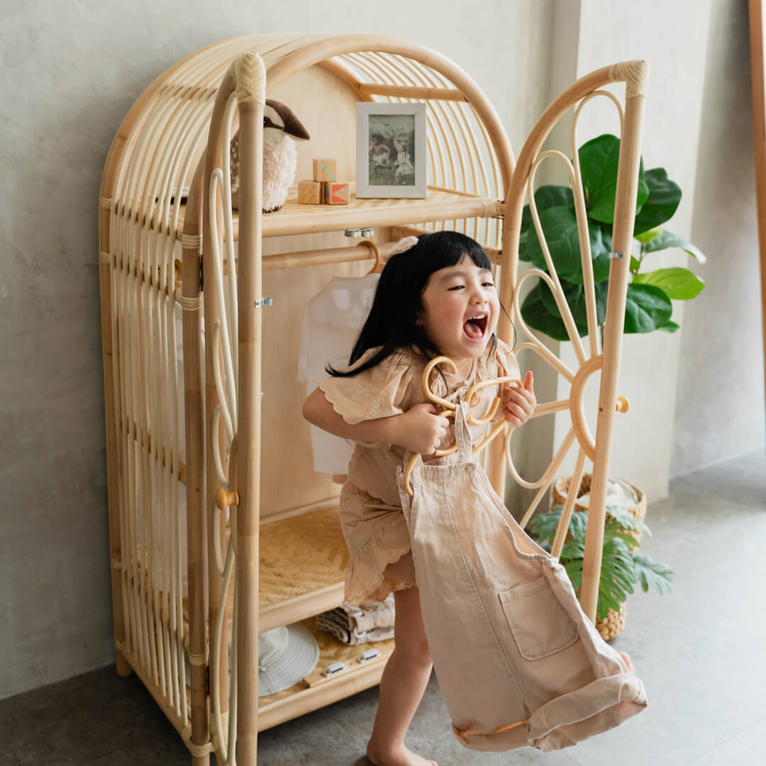 Happy child holding clothes in front of the Blossom Kids Wardrobe by MOMIJI, styled in a cozy room with indoor plants.