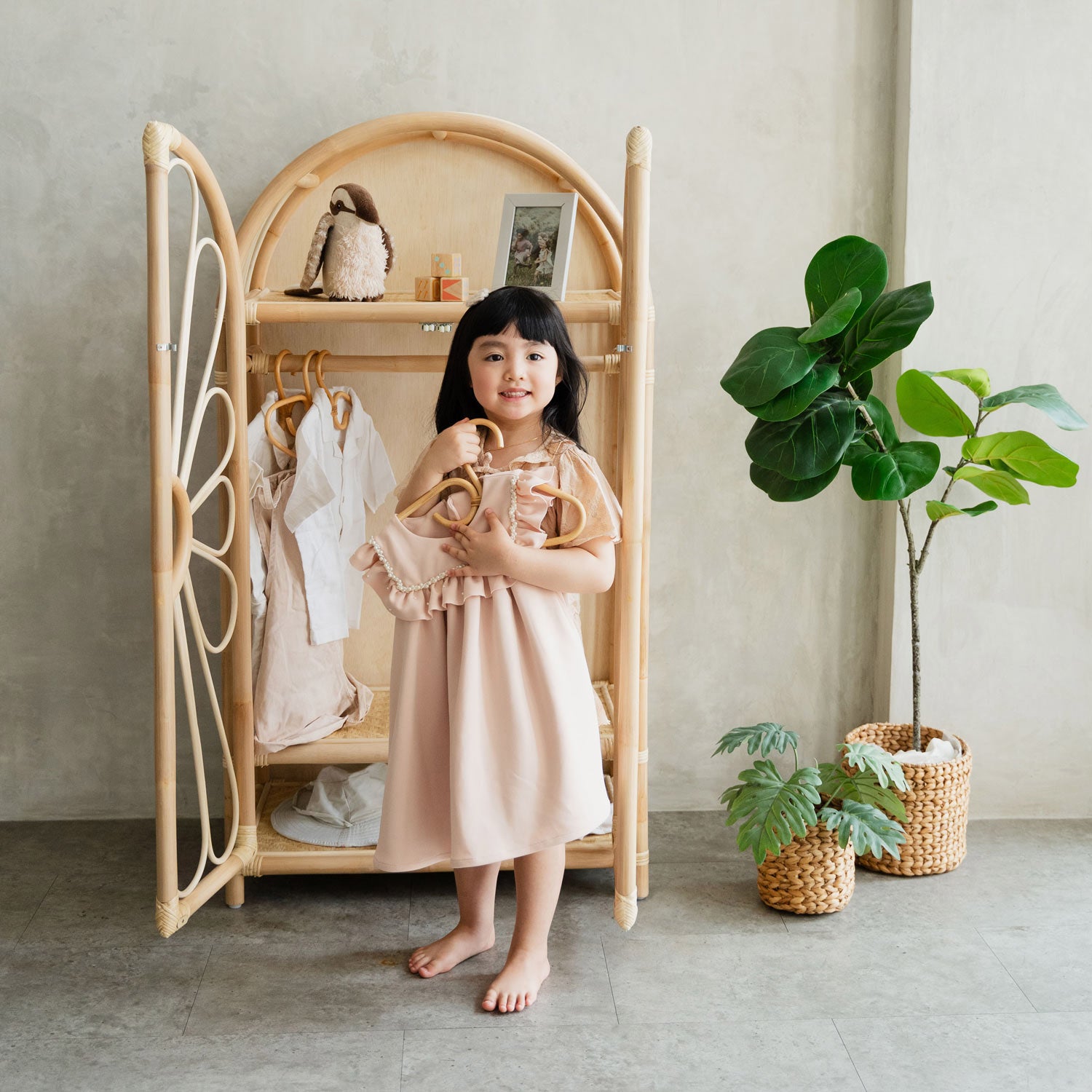Young girl in a pink dress standing next to a rattan wicker wardrobe cabinet with her clothes hanging on the rack
