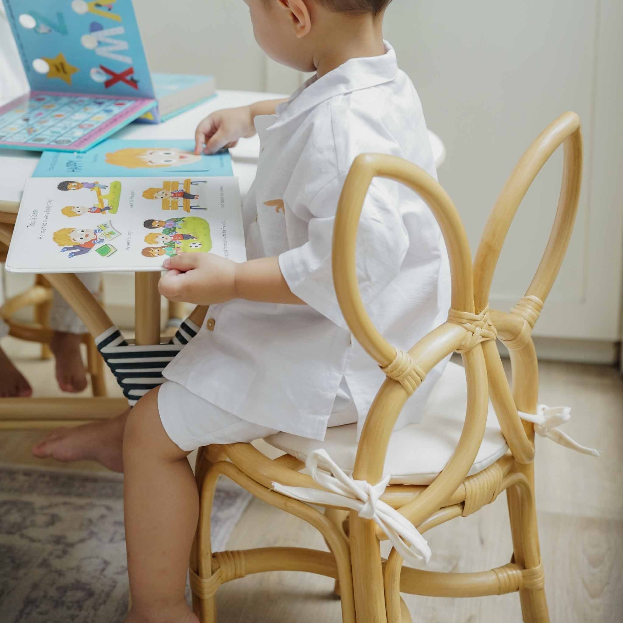 Back view of a child seated on a handcrafted natural rattan wicker Butterfly Kids Chair by MOMIJI, showcasing the butterfly-wing backrest and comfortable removable cushion while reading at a small table.