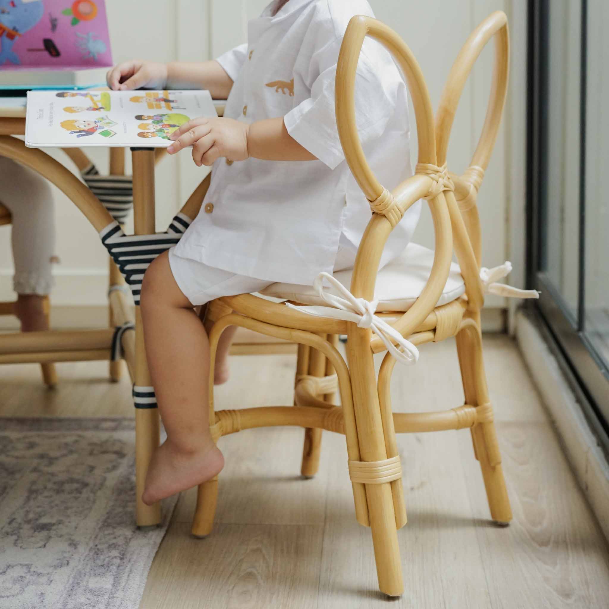 Back view of a child sitting on a handcrafted natural rattan wicker Butterfly Kids Chair by MOMIJI, featuring a butterfly-wing backrest and comfortable removable seat cushion, while reading at a small table.