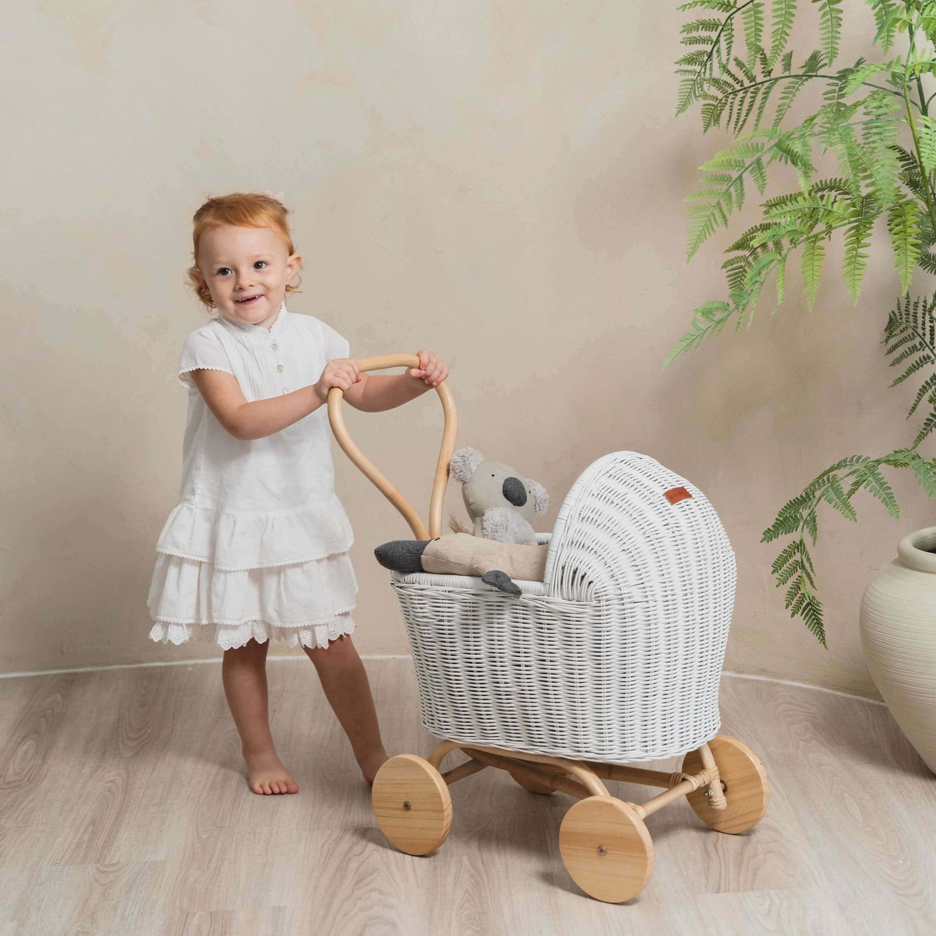 Child in a white dress pushing the Colette Doll Stroller by MOMIJI in white, styled in a room with a plant.
