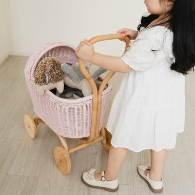 Child in a white dress pushing a handcrafted pink rattan wicker Colette Doll Stroller by MOMIJI on a wooden floor.