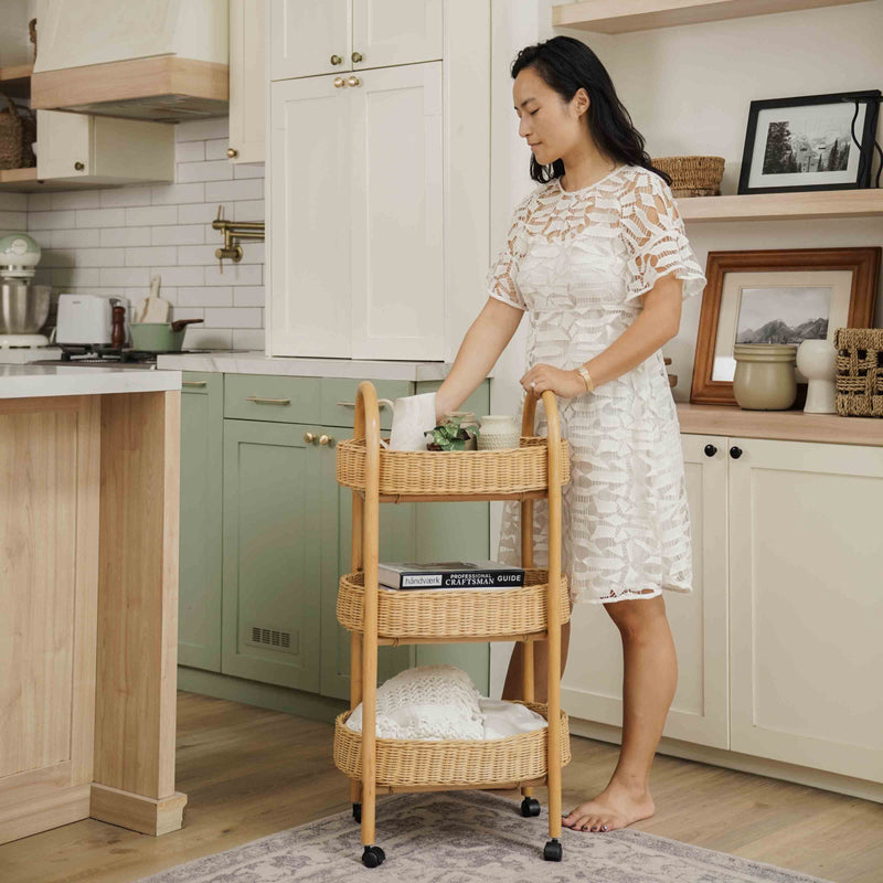 Woman standing in a kitchen with a handcrafted wicker rattan Con Craft Trolley by MOMIJI featuring three basket trays, wheels, and side handles