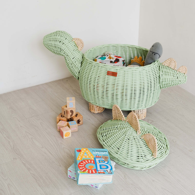 A mint green dinosaur rattan storage basket in large size by MOMIJI with toys inside the rattan basket, lid on the floor next to a pile of blocks and books on the floor.