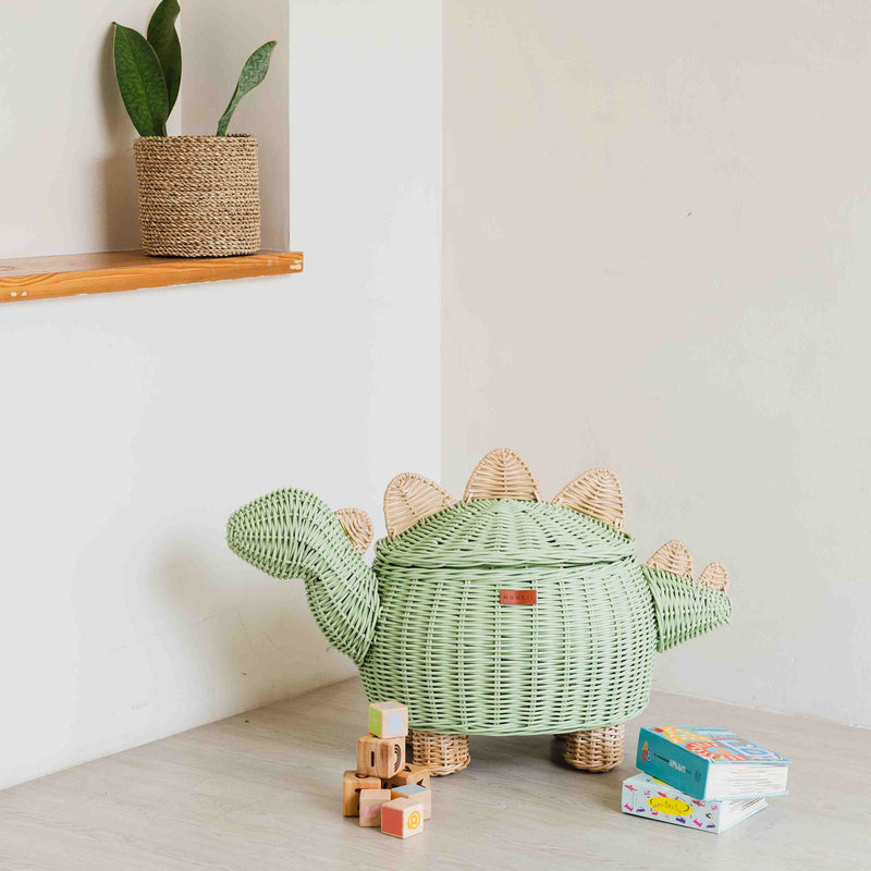 A mint green dinosaur rattan storage basket in large size by MOMIJI, placed on a floor next to a plant pot and children's blocks.
