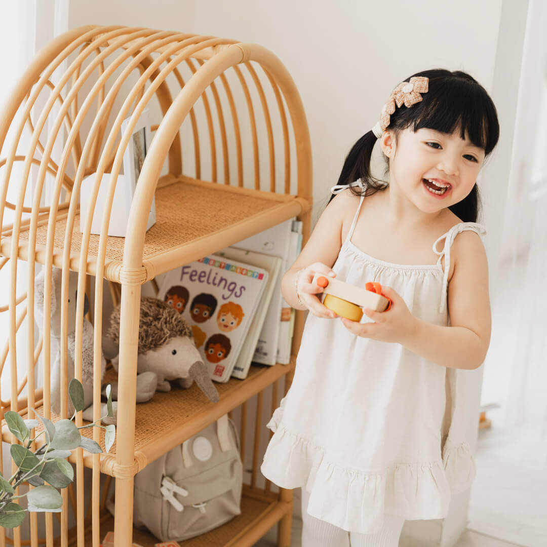 Smiling child holding a toy camera beside the Daisy Arch Shelf by MOMIJI in a cozy indoor setting.