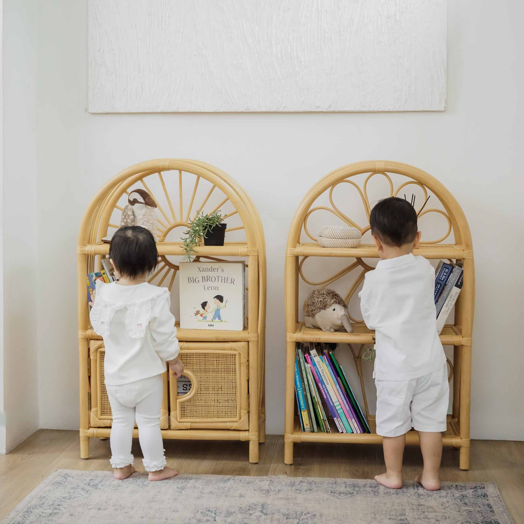 Two children in white outfits standing  in front of Daisy Shelves and Sunshine Arch Shelves by MOMIJI, placing books onto the shelves and showcasing the child-friendly design that encourages independent organisation and easy access to books and toys.