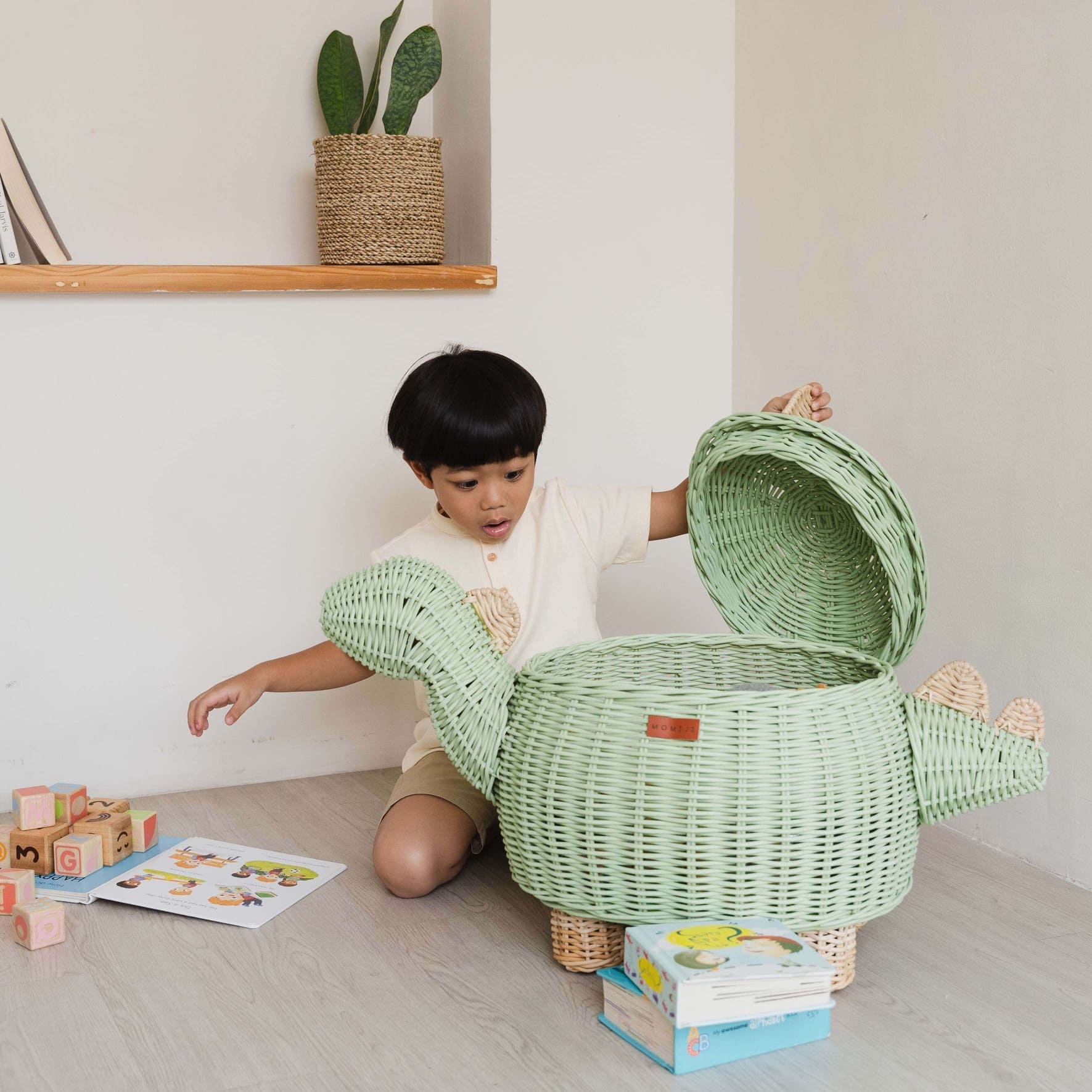 Child surprisingly opened his mint green dinosaur rattan storage basket in large size by MOMIJI with toys and books on the floor.