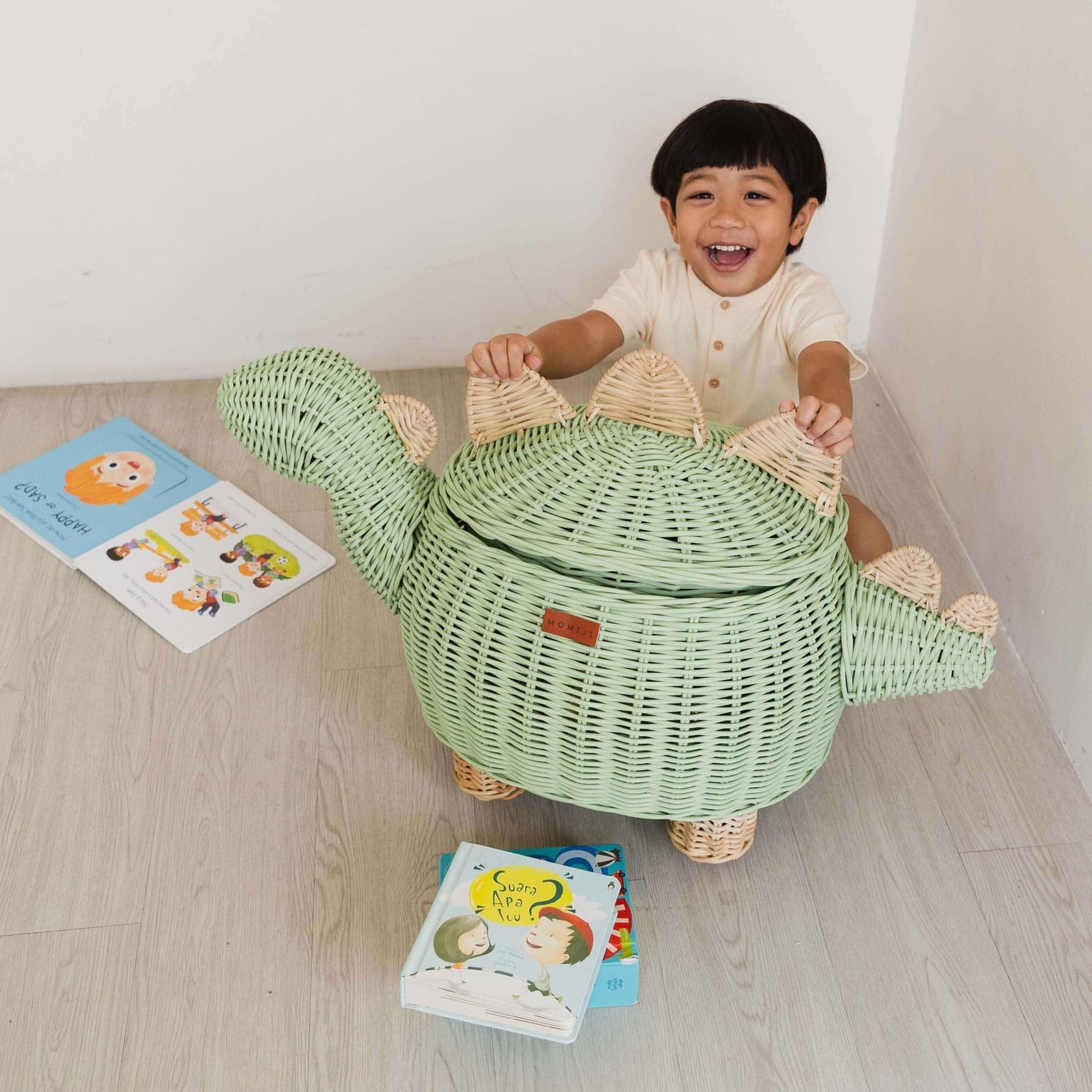 Child happily playing with his Mint Green Dinosaur Storage Rattan Basket by MOMIJI in extra large size, with books on the wooden floor.