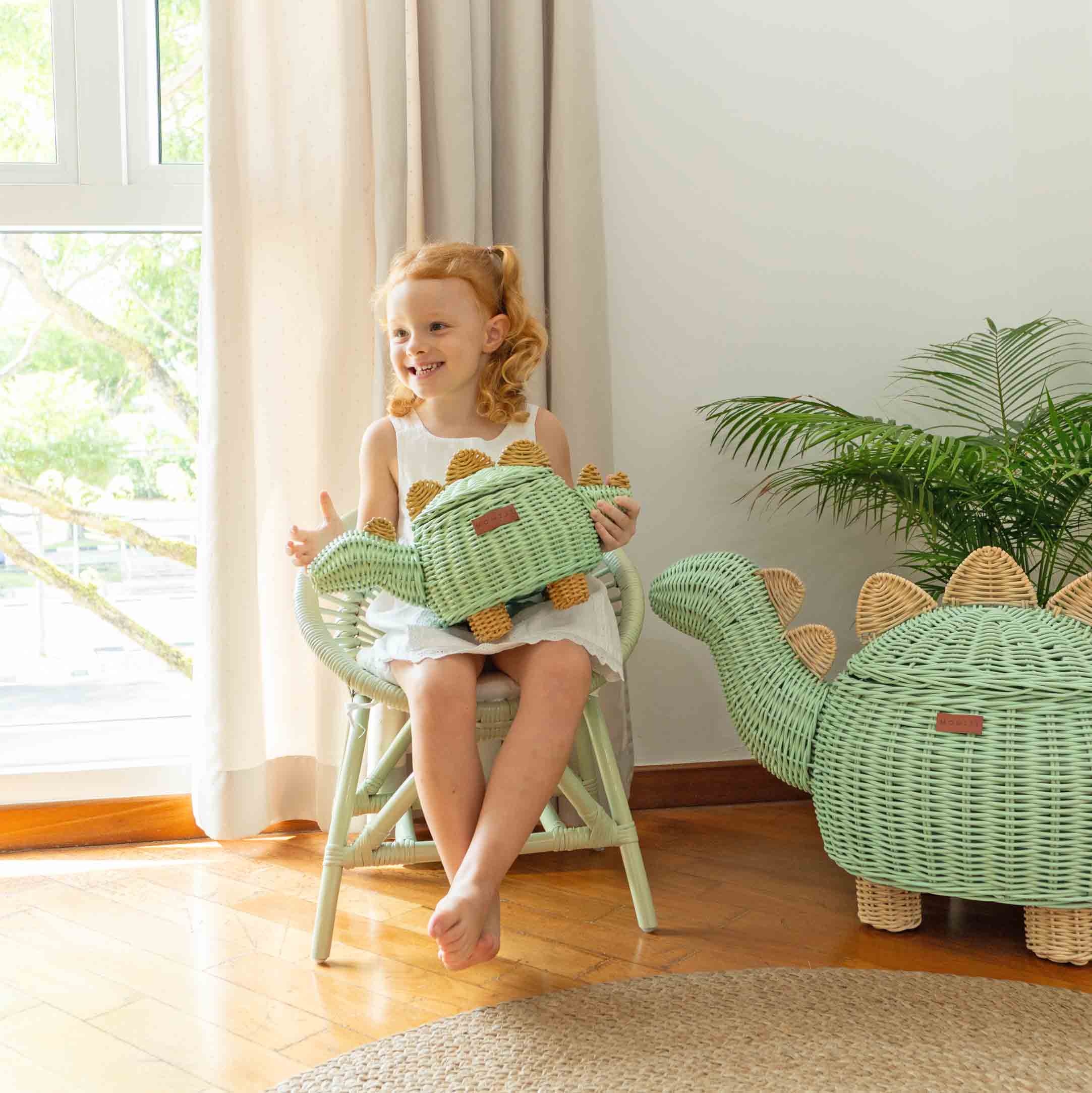 Child sitting on a chair holding a Dinosaur Rattan Storage Basket Small by MOMIJI in a bright room with a plant and an extra large Dinosaur Rattan Storage Basket on the floor.