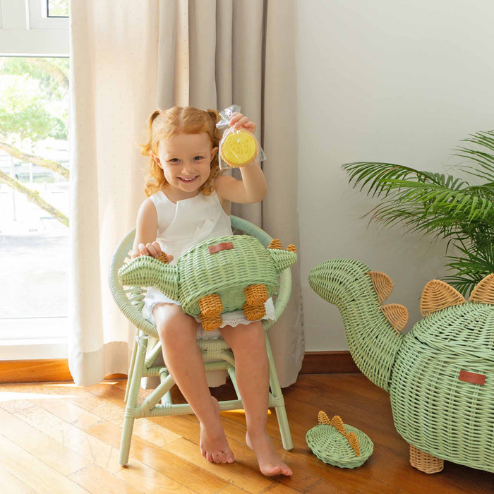 Child sitting on a green chair holding a Dinosaur Rattan Storage Basket in Small size by MOMIJI, with another dinosaur rattan basket on the floor.