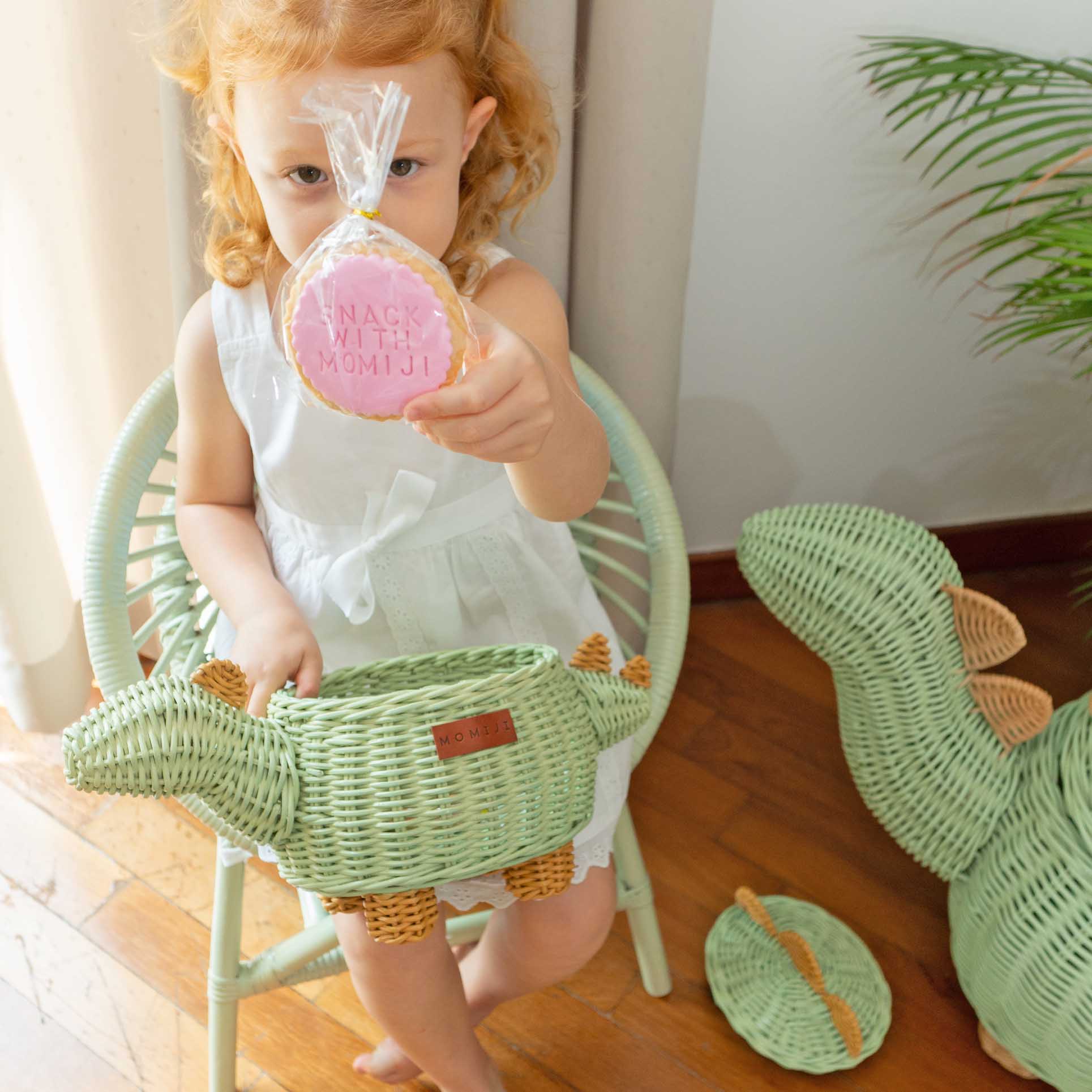 Child holding a cookie and a Dinosaur Rattan Storage Basket in Small size by MOMIJI sitting on a green chair with another dinosaur rattan basket on the floor.