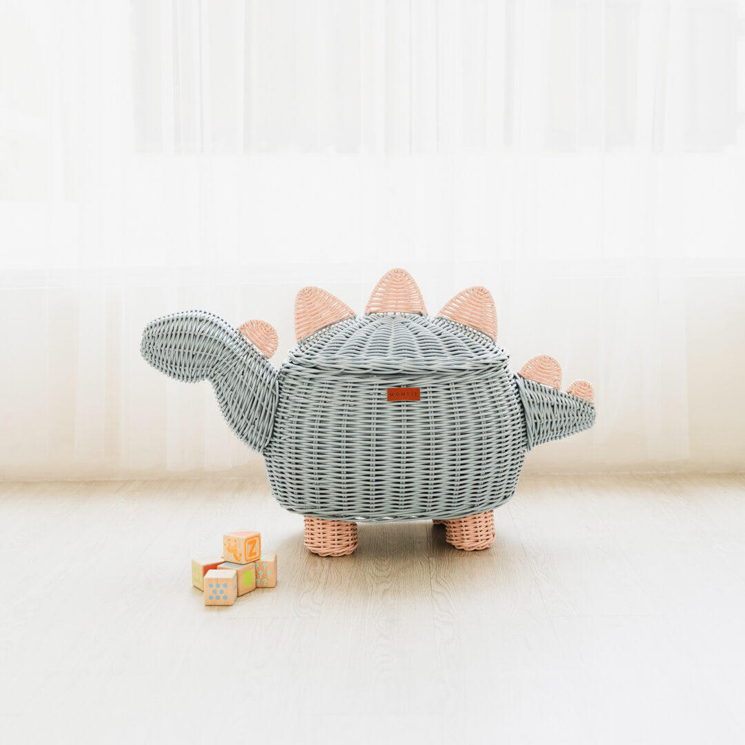 A pastel blue and pink dinosaur rattan storage basket by MOMIJI designed to resemble a stegosaurus, placed on a wooden floor next to some toy boxes.