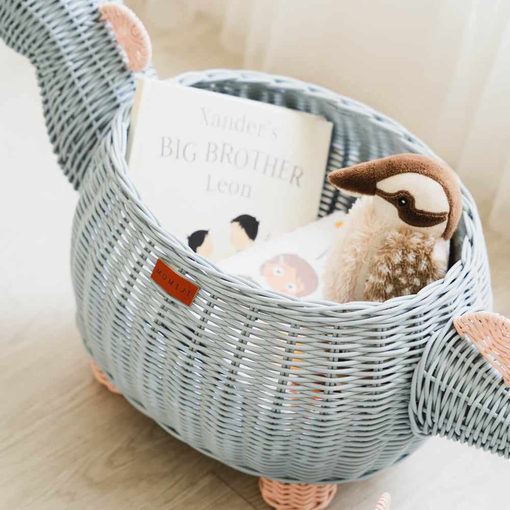 Close up shot inside pastel blue and pink dinosaur rattan storage basket by MOMIJI with a stuffed toy and books inside the rattan basket.