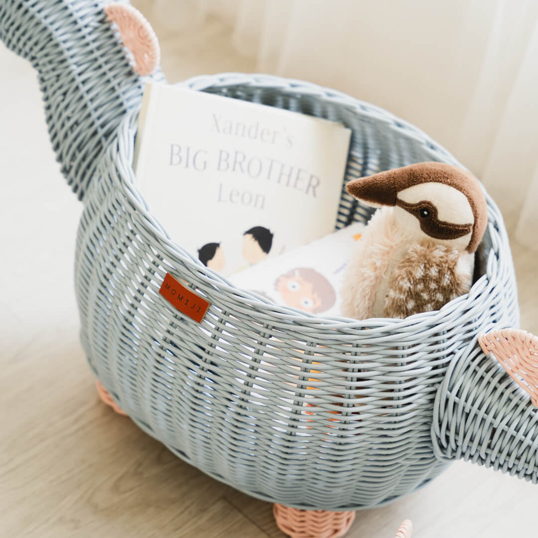Close up shot inside pastel blue and pink dinosaur rattan storage basket by MOMIJI with a stuffed toy and books inside the rattan basket.