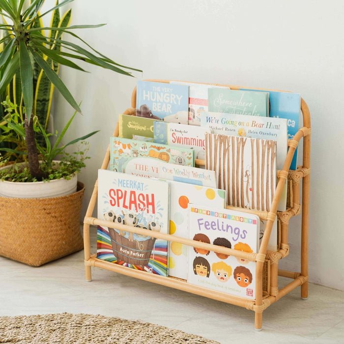 Children’s books on the handwoven rattan wicker Elena Kids Montessori Bookshelf – Natural by MOMIJI in a room with a plant and rug.