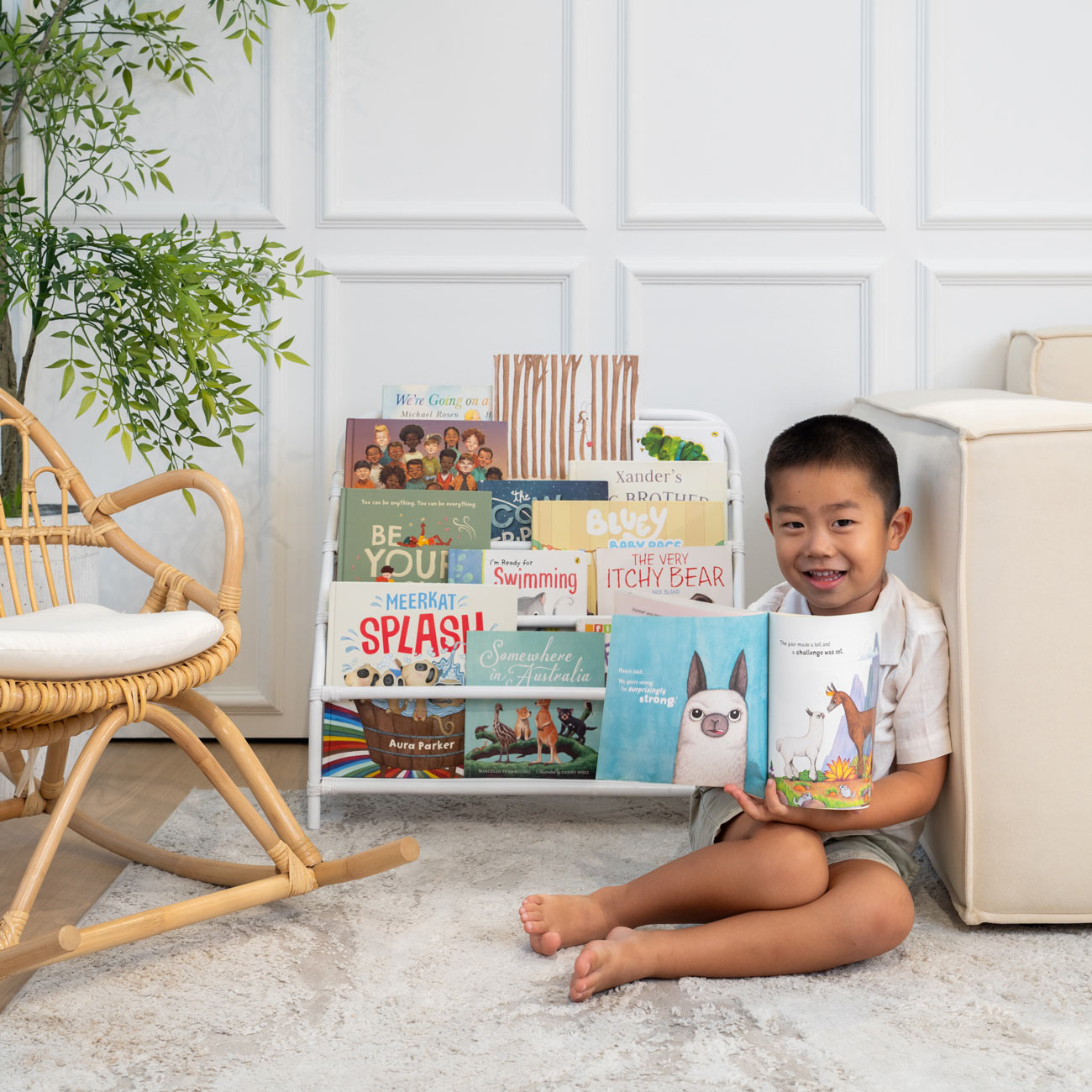 Smiling child sitting on the floor holding a book in front of the handwoven rattan wicker Elena Kids Montessori Bookshelf – White by MOMIJI.