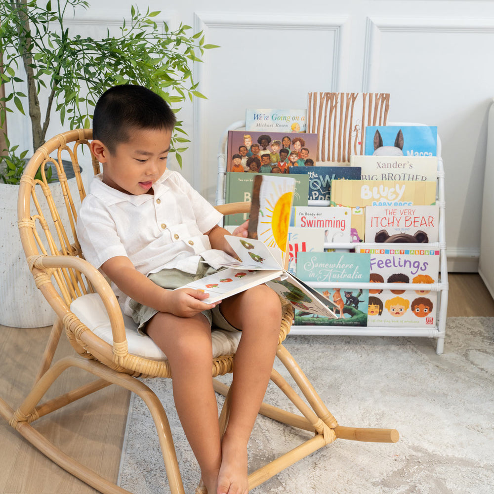 Child sitting on a rattan chair reading a book, with the handwoven rattan wicker Elena Kids Montessori Bookshelf – White by MOMIJI in the background.