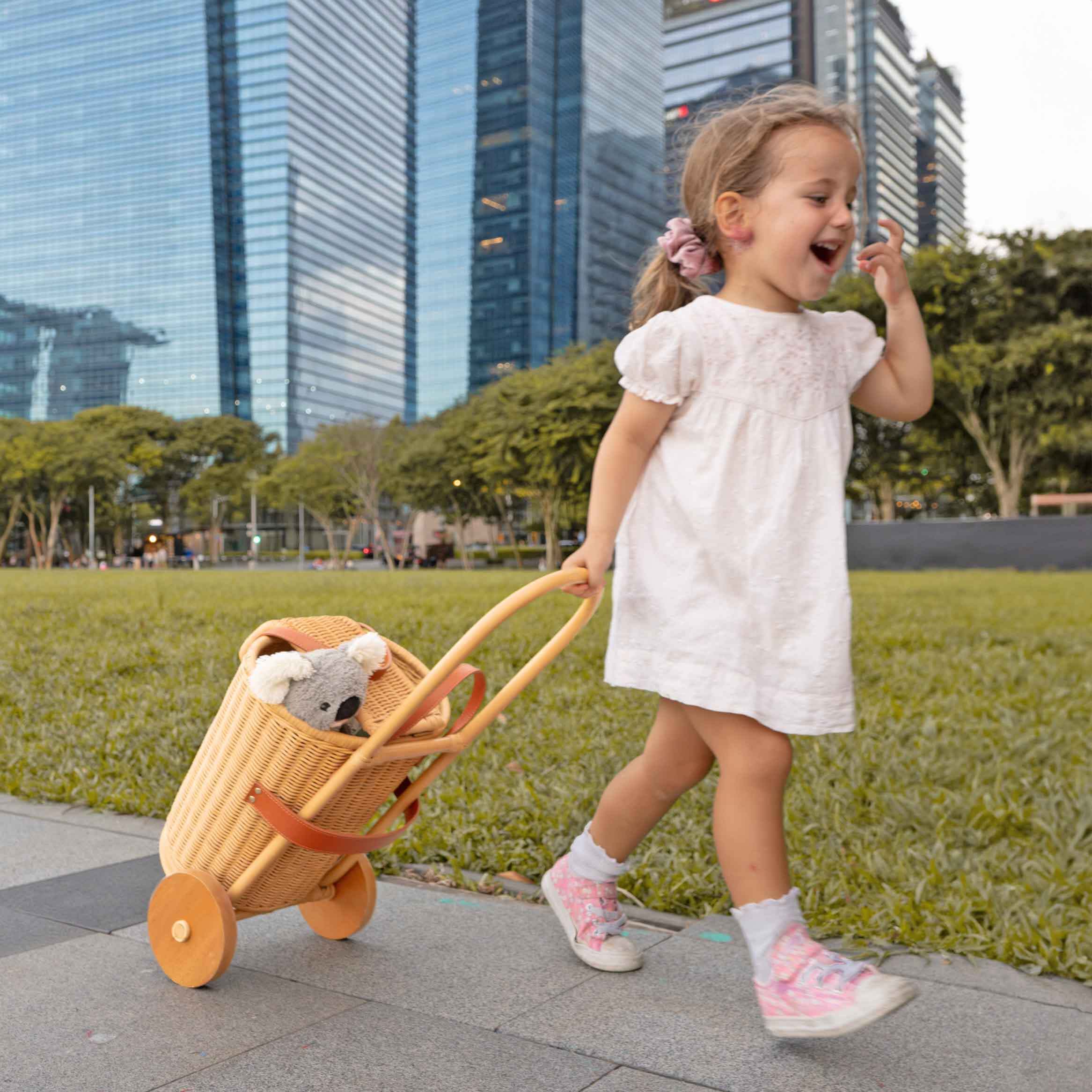 Child pulling the Eric Kids Shopping Trolley by MOMIJI filled with dolls in an outdoor park with buildings in the background.