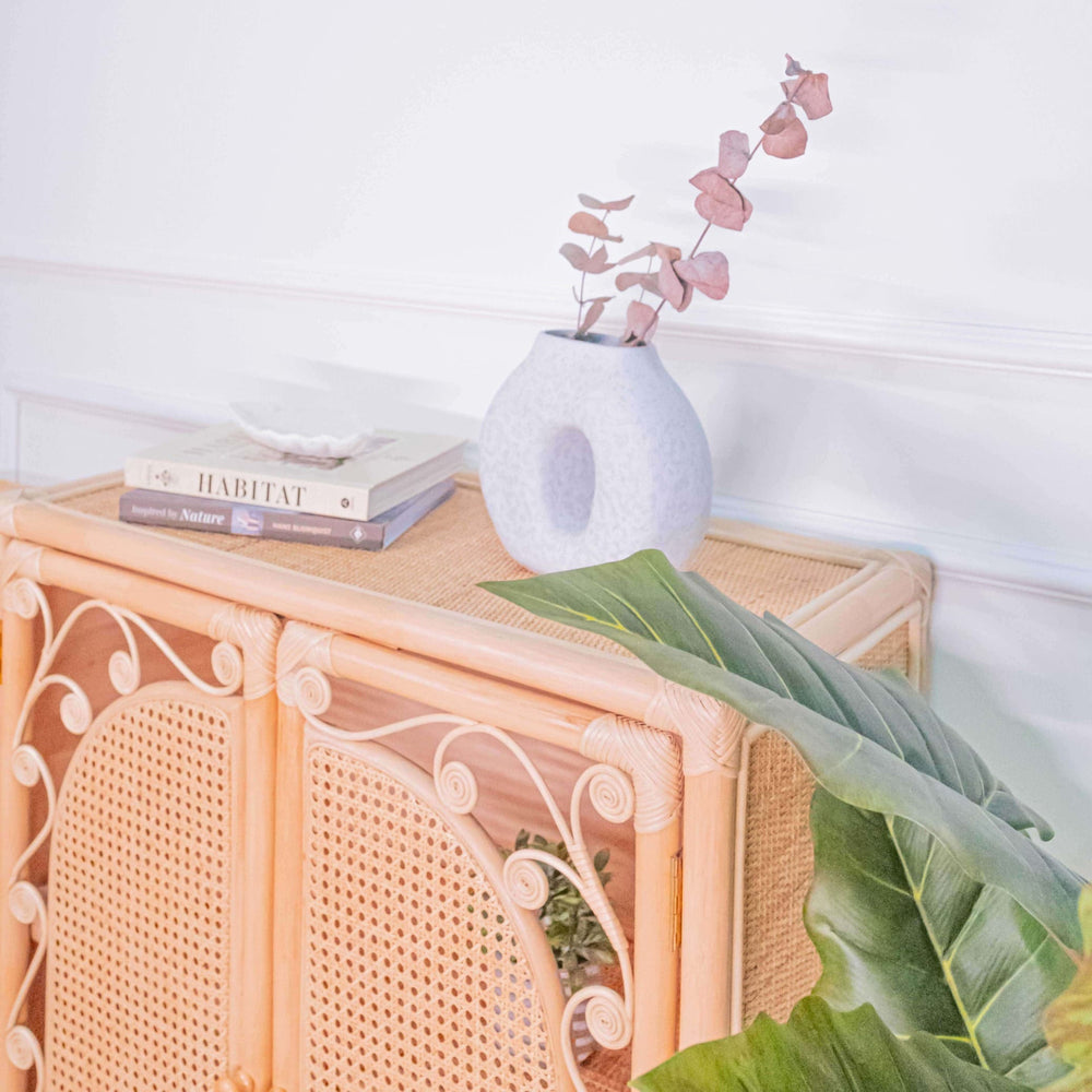 Top of the handcrafted rattan wicker Fern Storage Cabinet by MOMIJI styled with a vase and books in a room with plants.