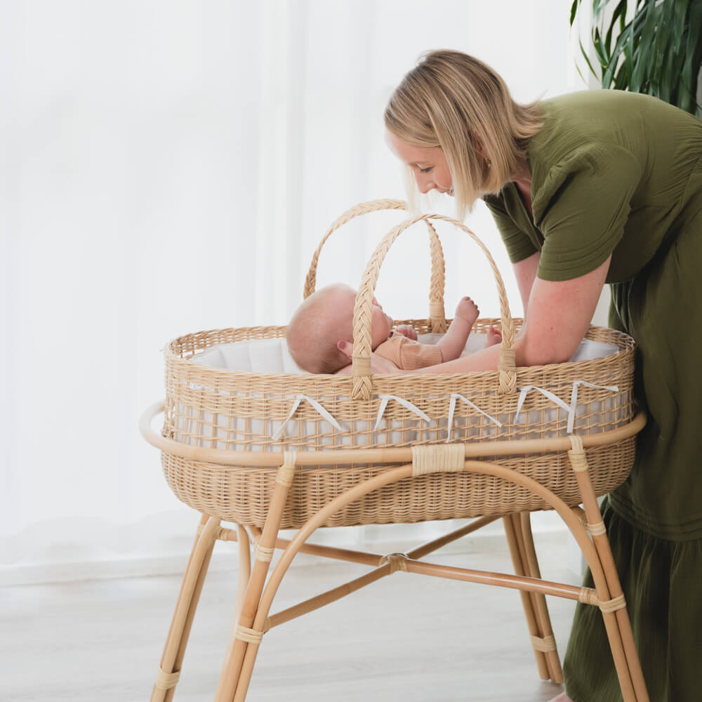 Woman interacting with a baby resting in a wicker Moses basket on a fixed stand by MOMIJI.