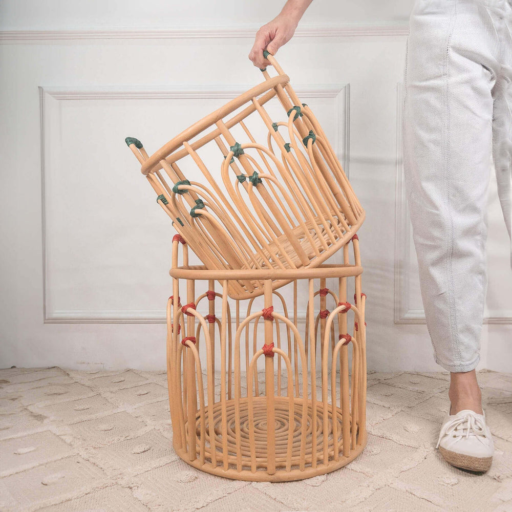Woman placing the smaller handcrafted rattan wicker Floret Basket with green binding into the larger basket with red binding by MOMIJI, styled on a rug.