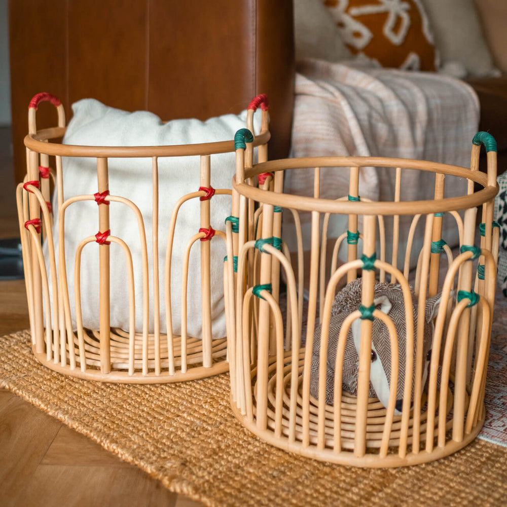 Large and small handcrafted rattan wicker Floret Baskets by MOMIJI placed side by side near a sofa, in natural rattan with green and red bindings.