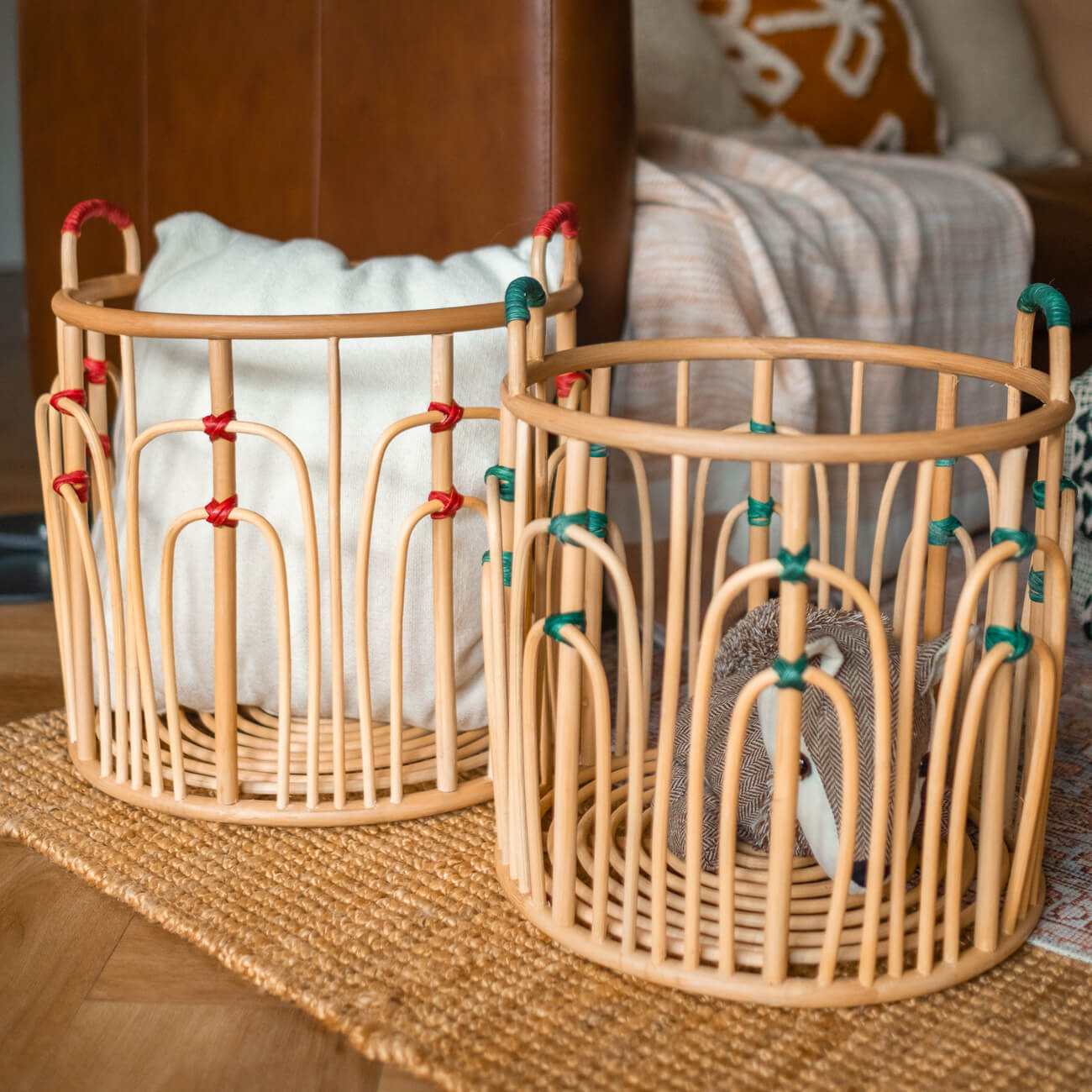 Large and small handcrafted rattan wicker Floret Baskets by MOMIJI placed side by side near a sofa, in natural rattan with green and red bindings.