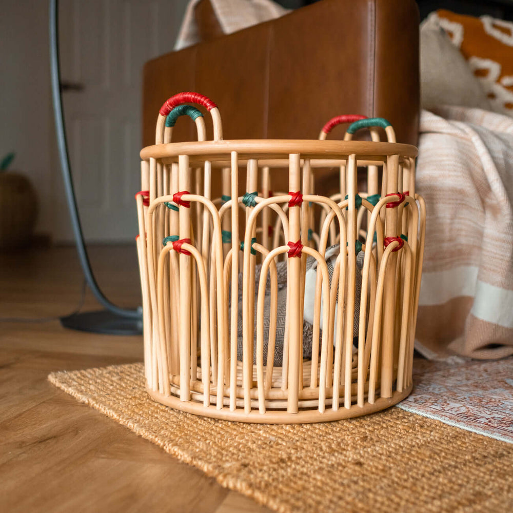 Stacked handcrafted rattan wicker Floret baskets by MOMIJI filled with dolls on a rug indoors in natural rattan with green and red bindings.