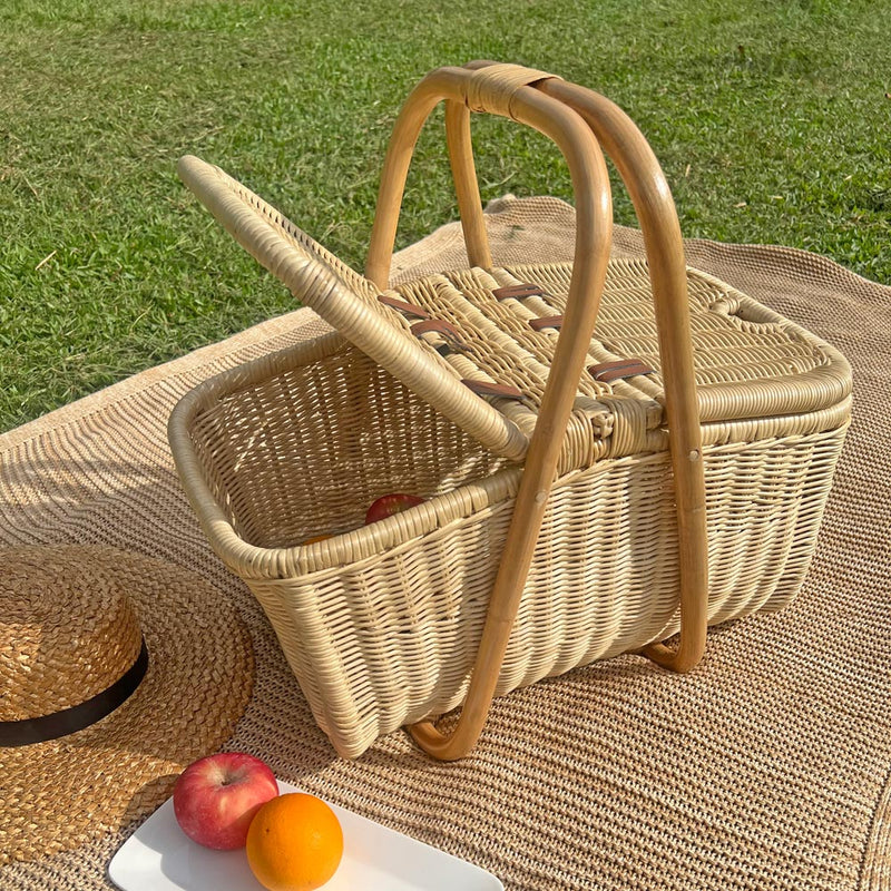 Aurelie rattan picnic basket by MOMIJI on a blanket with a straw hat and fruits on a grassy background.