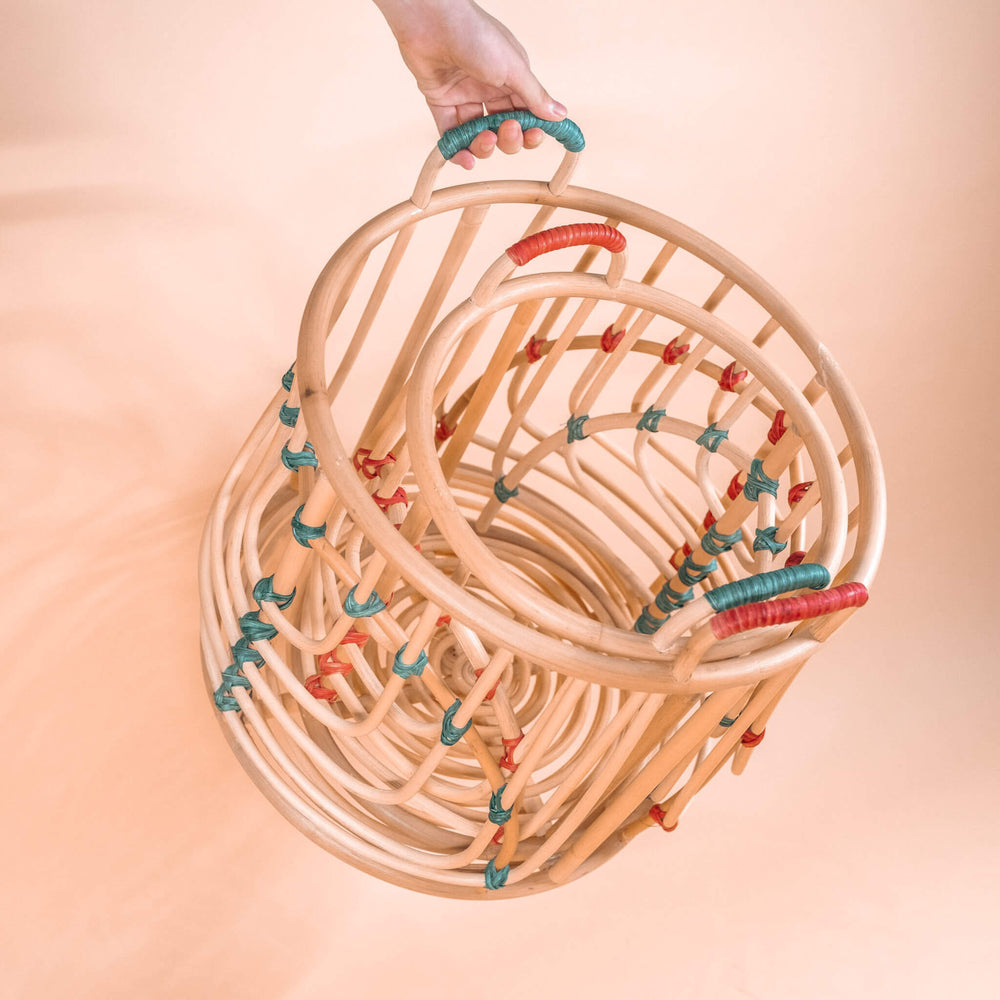 Hand holding the handcrafted Iris baskets set by MOMIJI in natural rattan with red and green bindings stacked together, shown in an angled view.